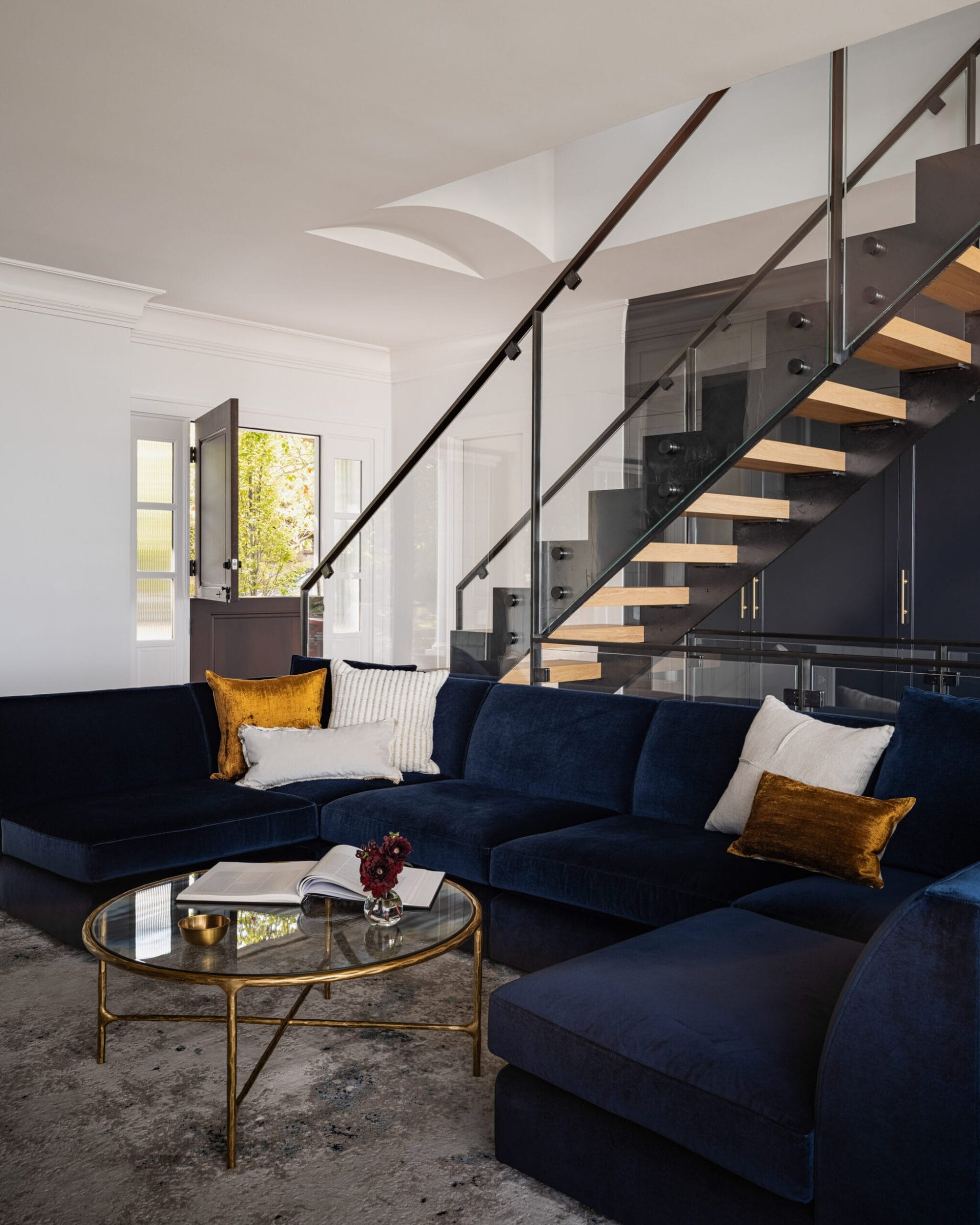 Modern living room with a dark blue sectional sofa, gold-accented round glass coffee table, and a staircase with glass railings. Decorative cushions accent the seating area.