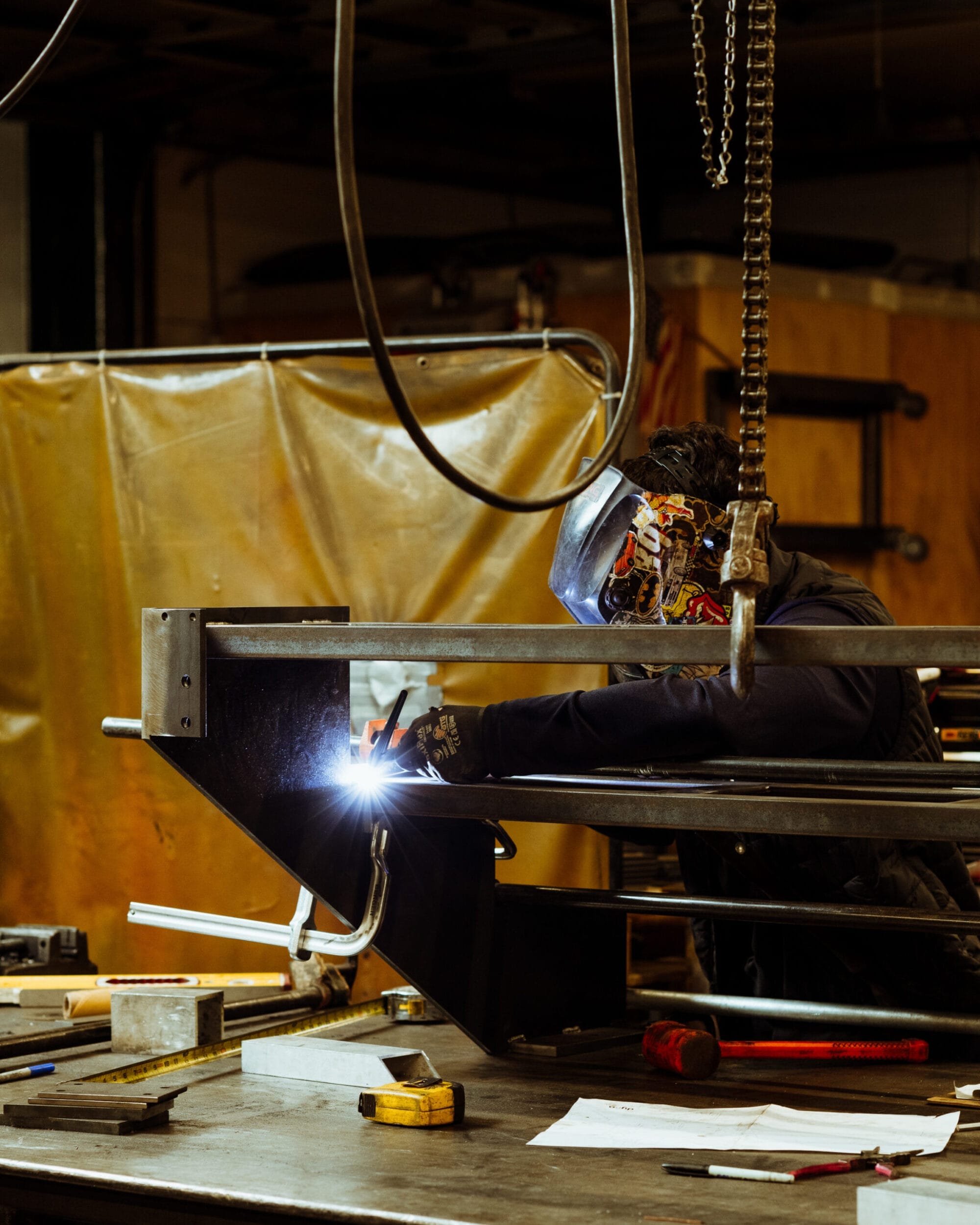 A skilled person is welding metal pieces at a workbench in Dyna Construction's industrial workshop, donning a protective helmet and gloves, ensuring every detail meets the high standards expected from a leading home builder in Seattle.