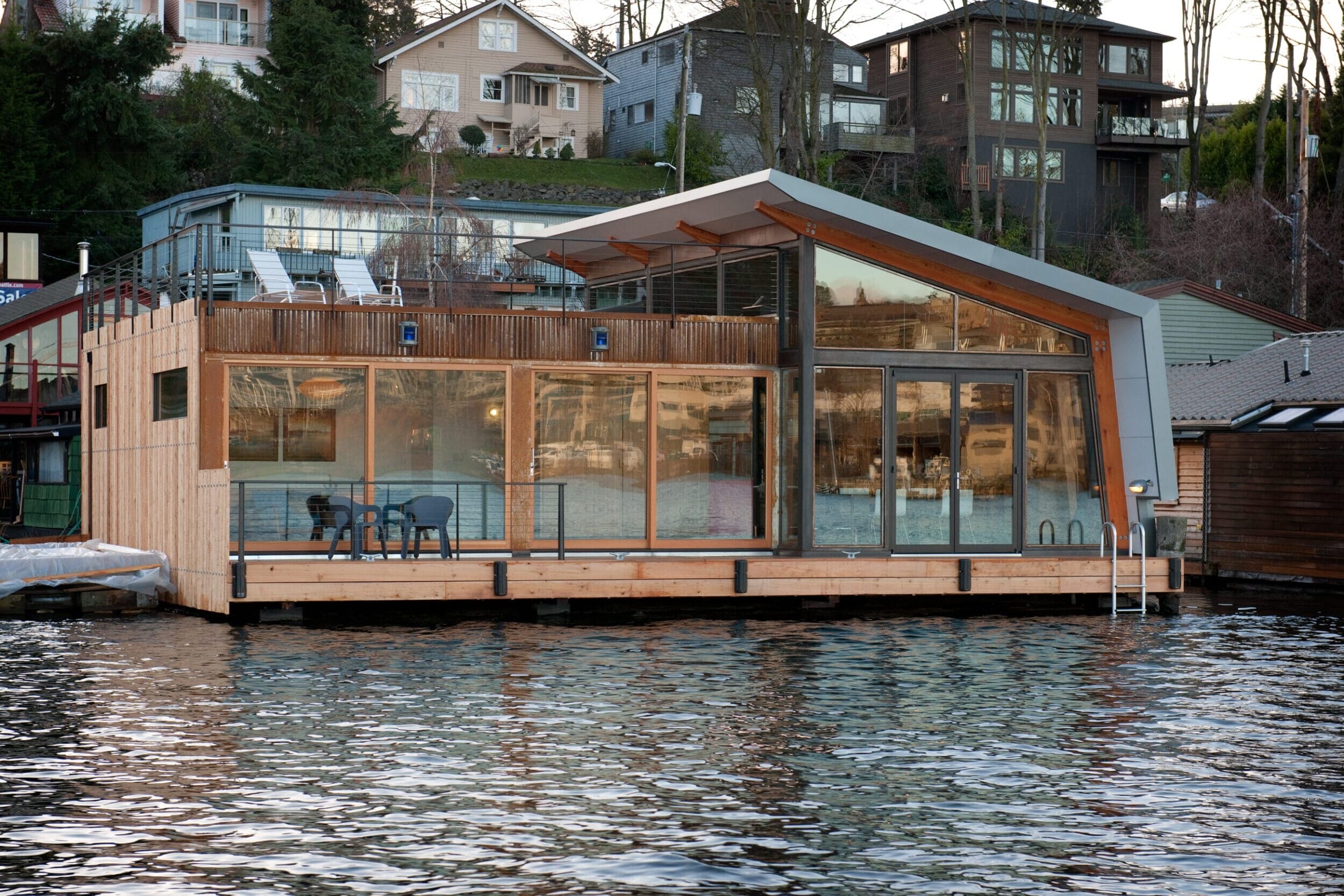 A modern floating house with large glass windows and a patio with chairs on a calm body of water, surrounded by trees and houses on a hill.