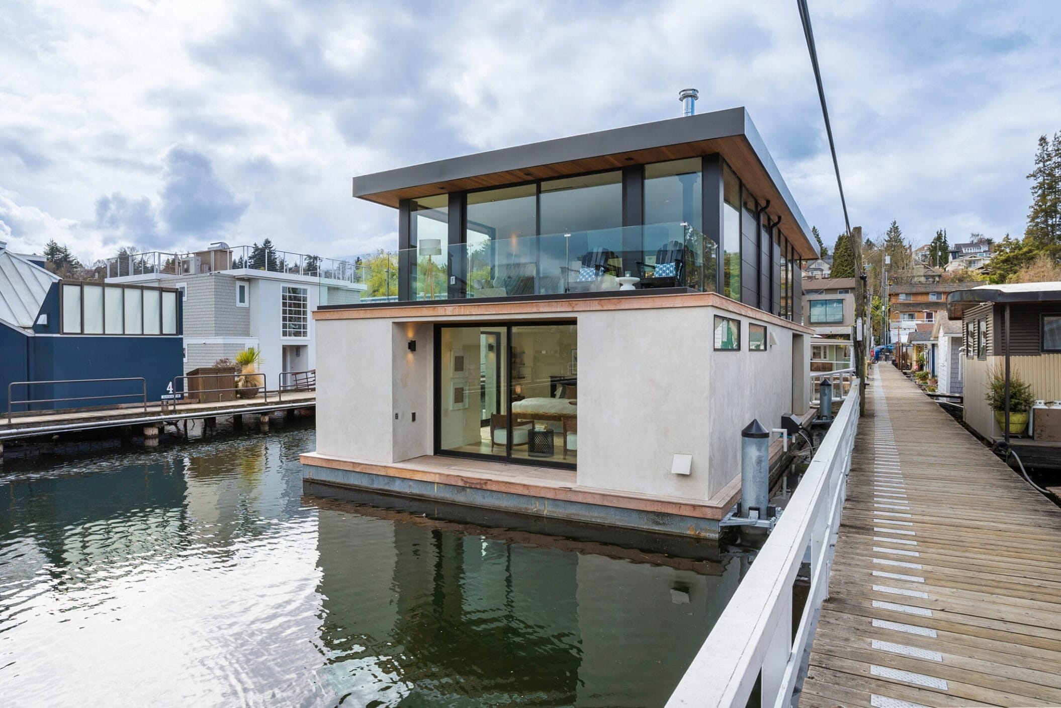 Modern houseboat with large windows and a flat roof, moored along a wooden walkway on a calm waterway, surrounded by other houseboats and trees under a cloudy sky.