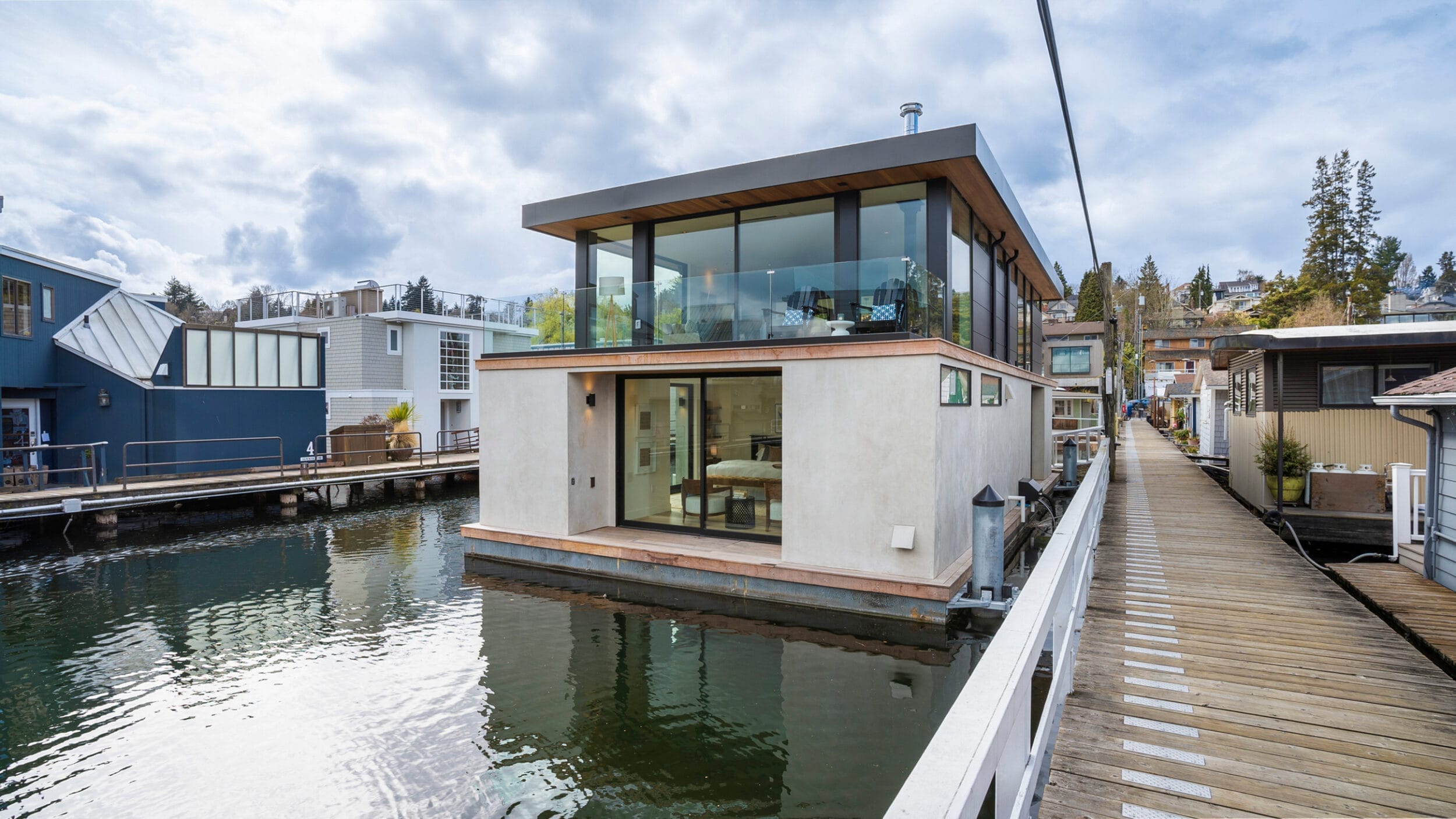 Modern houseboat with large windows and a flat roof, moored along a wooden walkway on a calm waterway, surrounded by other houseboats and trees under a cloudy sky.