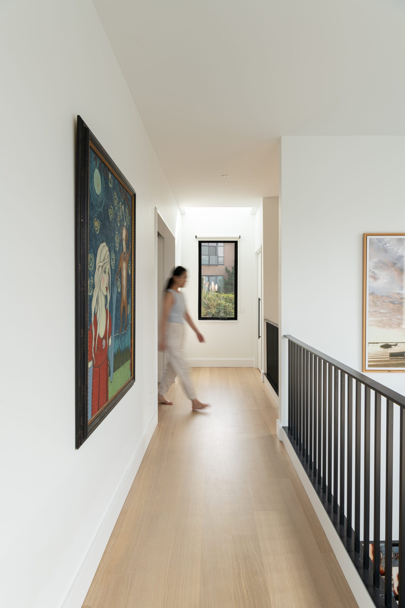 A person walks down a hallway with beige wooden flooring. Walls are white with artwork hanging. A railing is on the right, and a window with a view of trees is ahead.