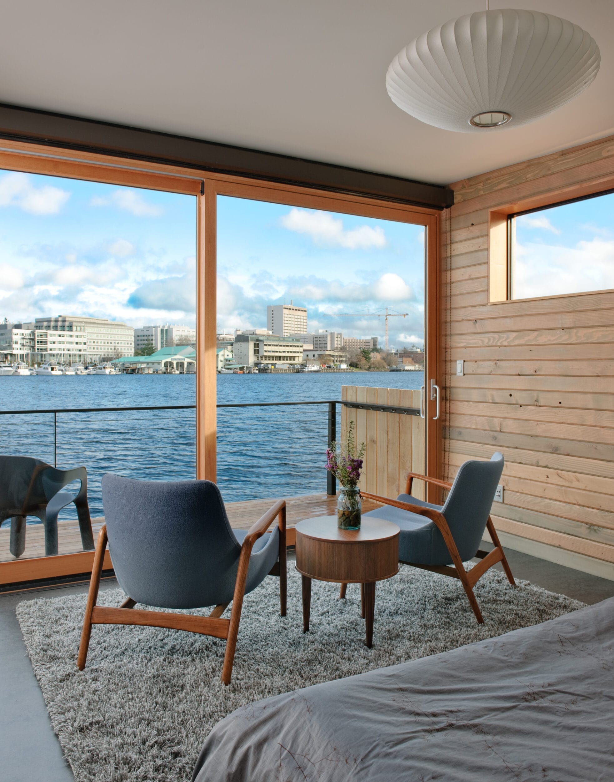 A modern room with a wooden interior, featuring two blue chairs and a small table on a rug, overlooking a waterfront view through large glass windows.