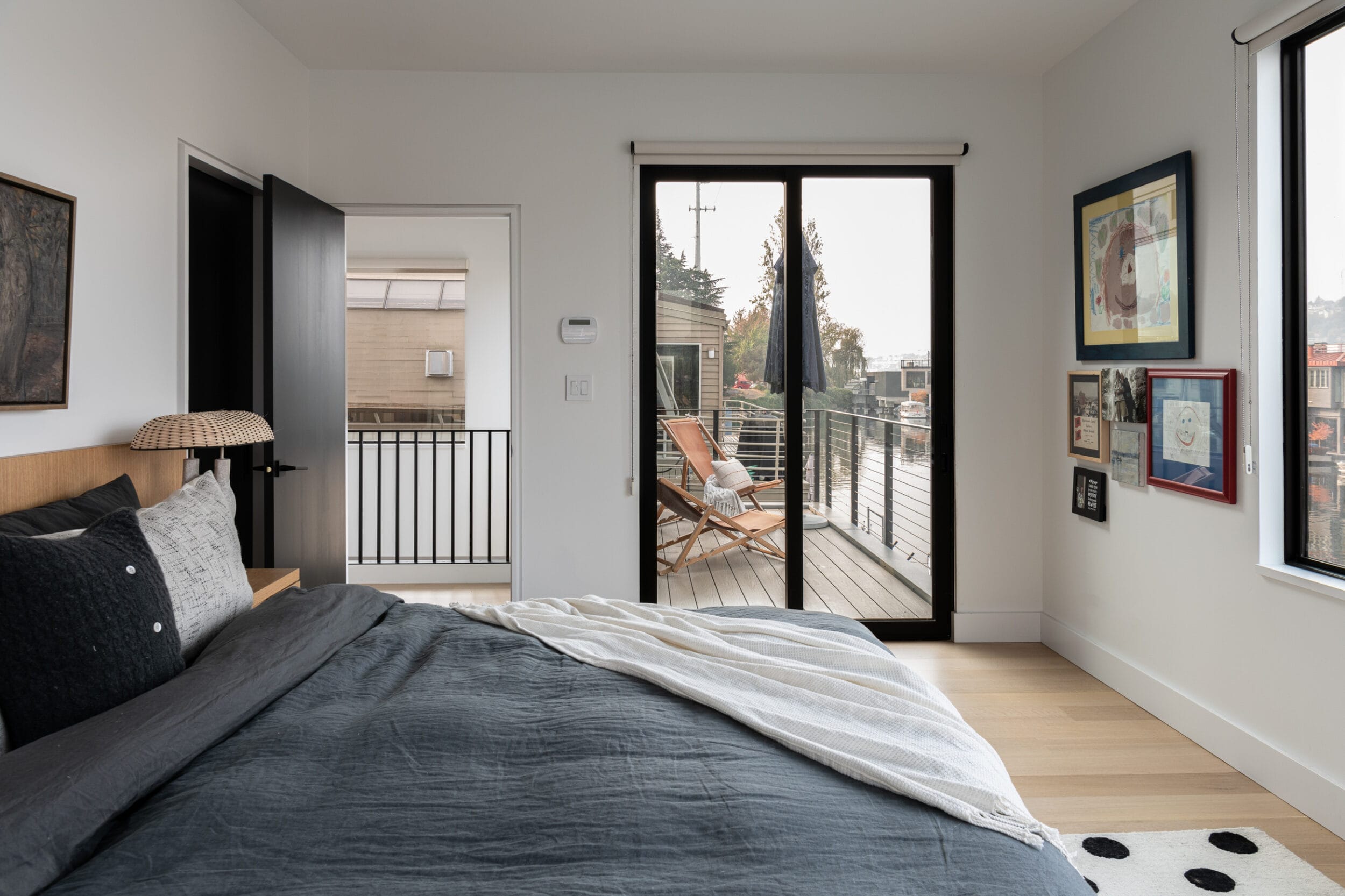 A modern bedroom with a double bed, gray bedding, and framed art on the walls. Glass doors open to a balcony with a chair. Light wood flooring and white walls create a minimalist look.