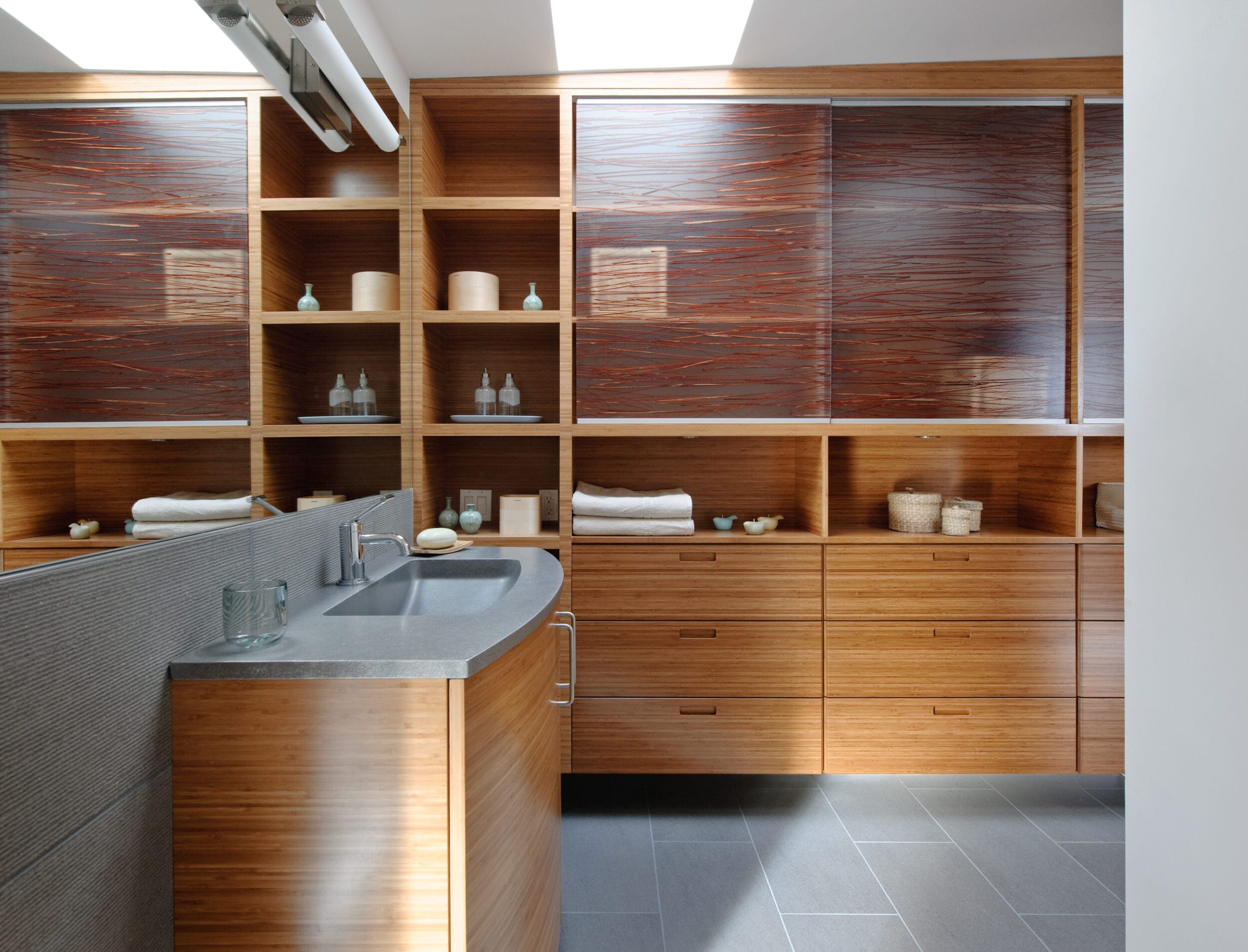 Modern bathroom with wood cabinetry, a gray countertop with a sink, open shelves with towels and toiletries, and a tiled floor. Natural light enters through a skylight.