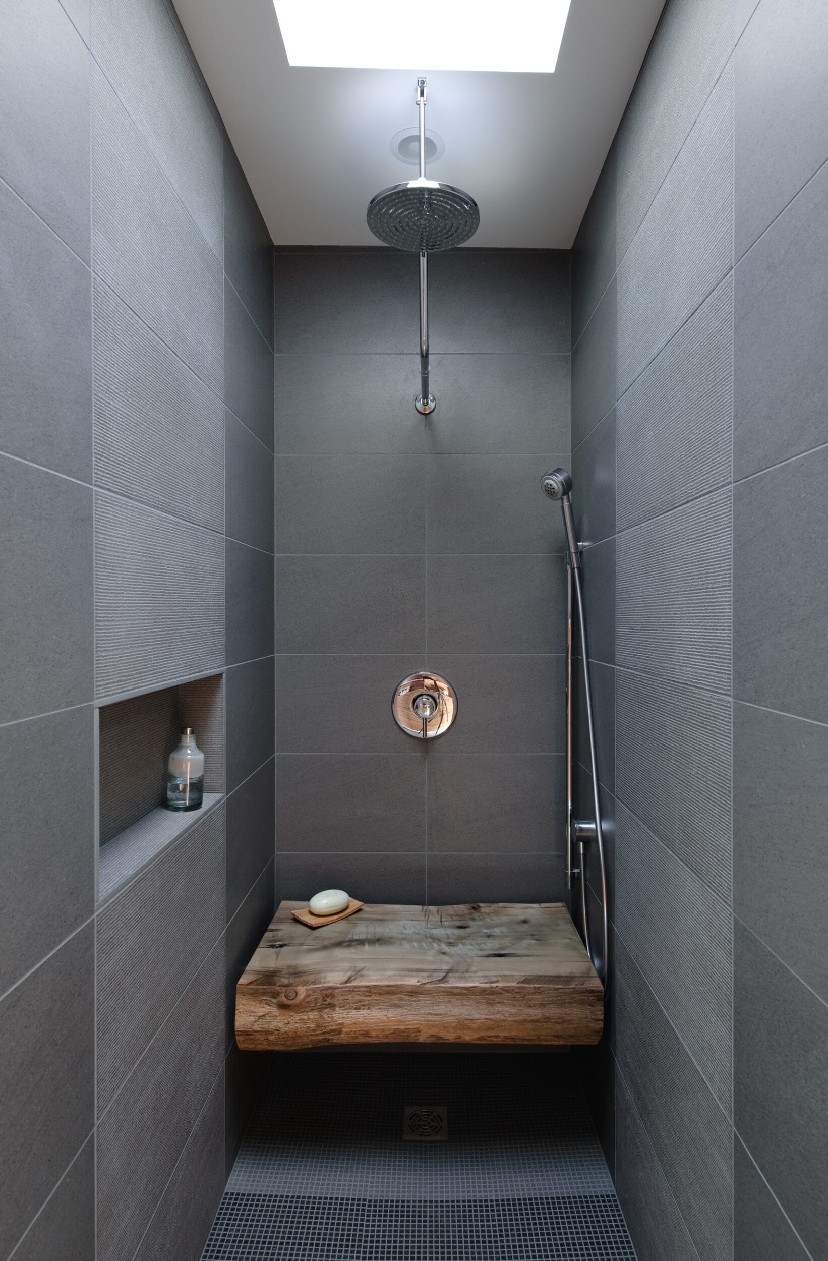 A minimalist shower with gray tiles, a stone bench, a rainfall showerhead, and a handheld nozzle. A soap dish and a bottle are on a recessed shelf on the left.