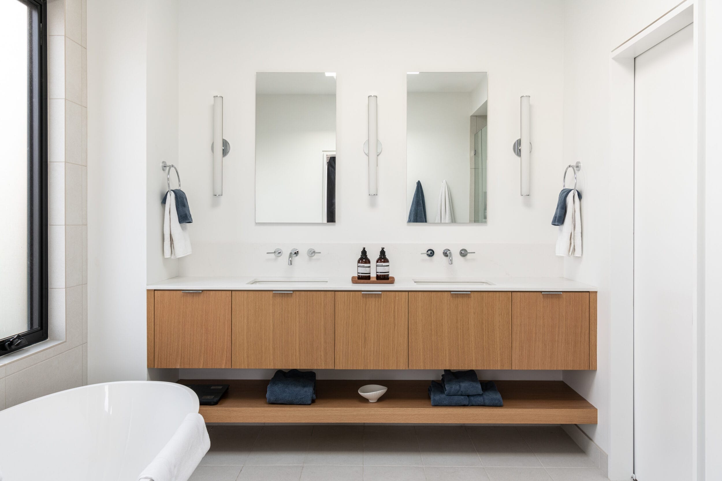 Minimalist bathroom with wooden double vanity, two mirrors, wall-mounted faucets, towels, and toiletries. A bathtub is on the left, and a modern light fixture is above each mirror.