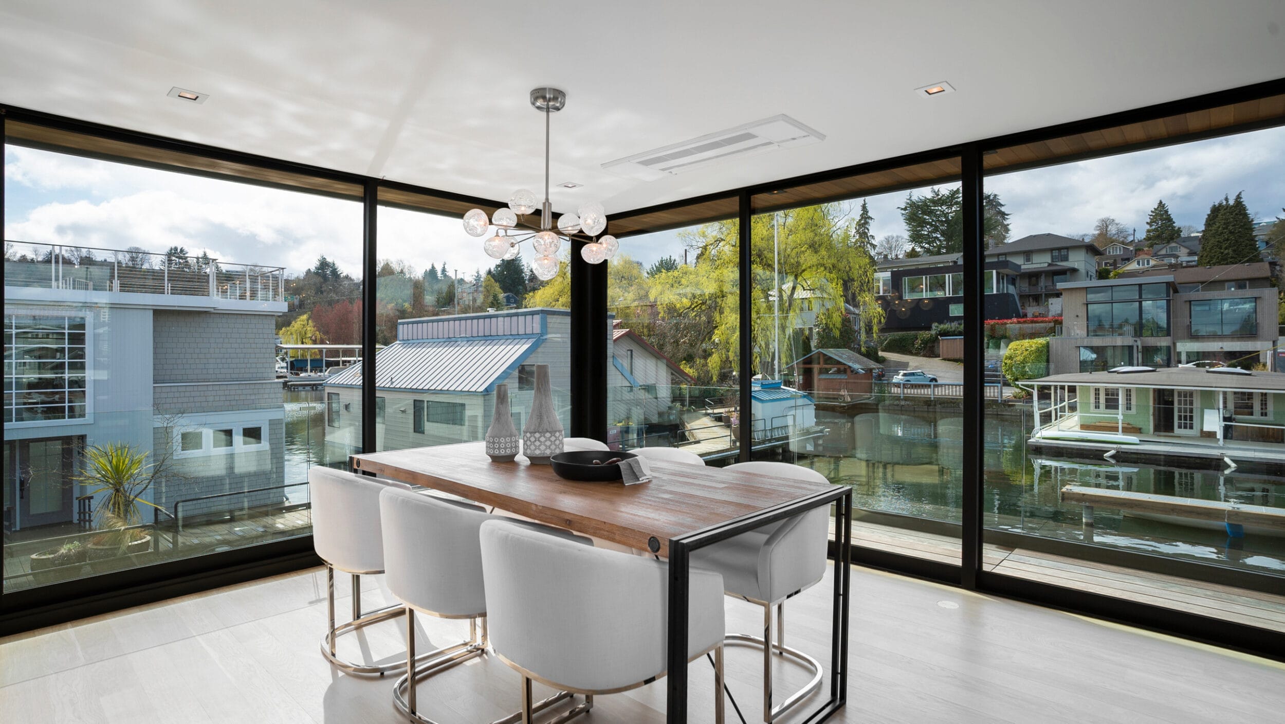 A modern dining area with a wooden table and white chairs surrounded by large glass windows overlooking a waterfront view with houses and boats.