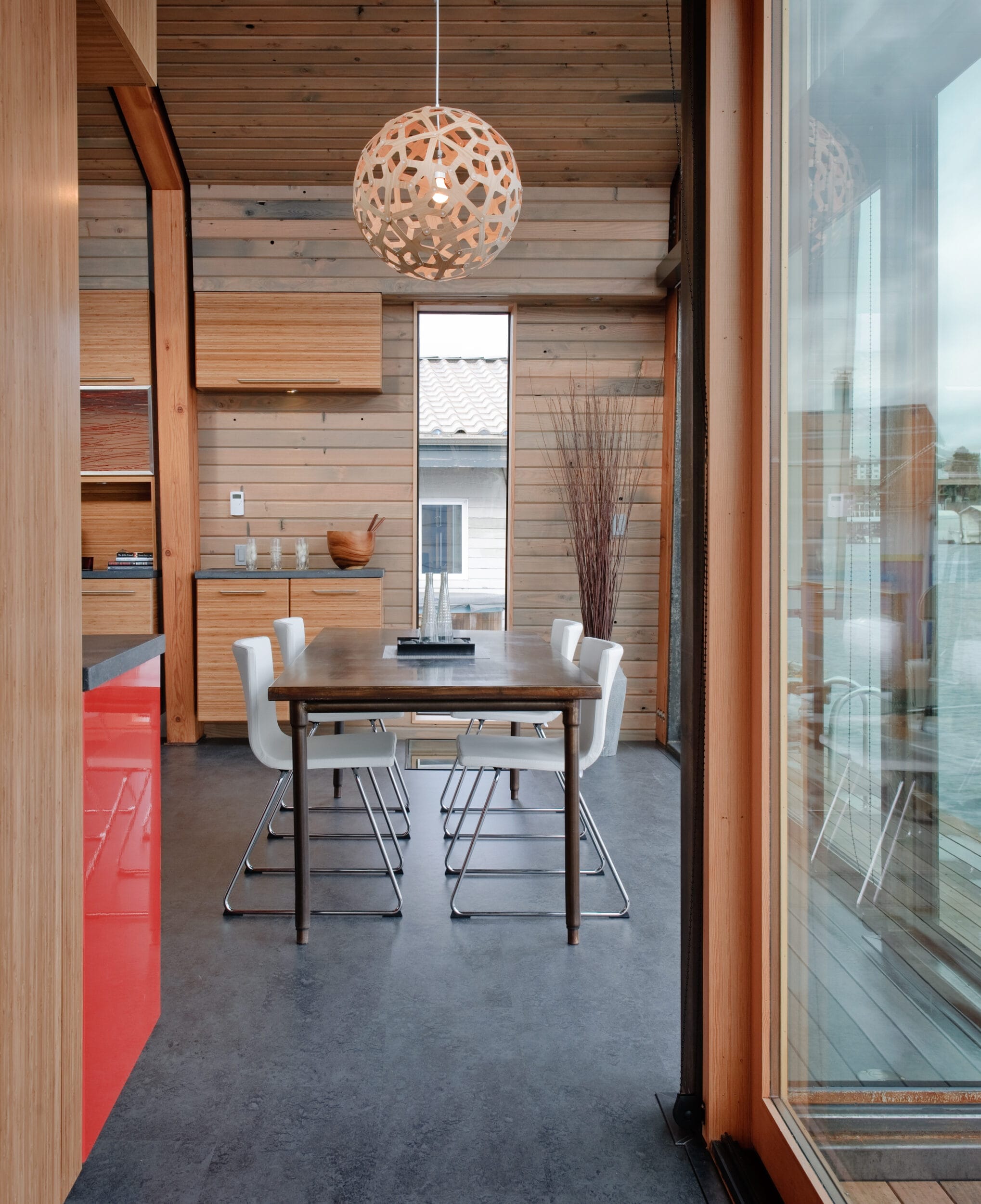 A modern dining area with a wooden table, four white chairs, and a decorative pendant light. The room features wood-paneled walls, large windows, and minimalistic decor.