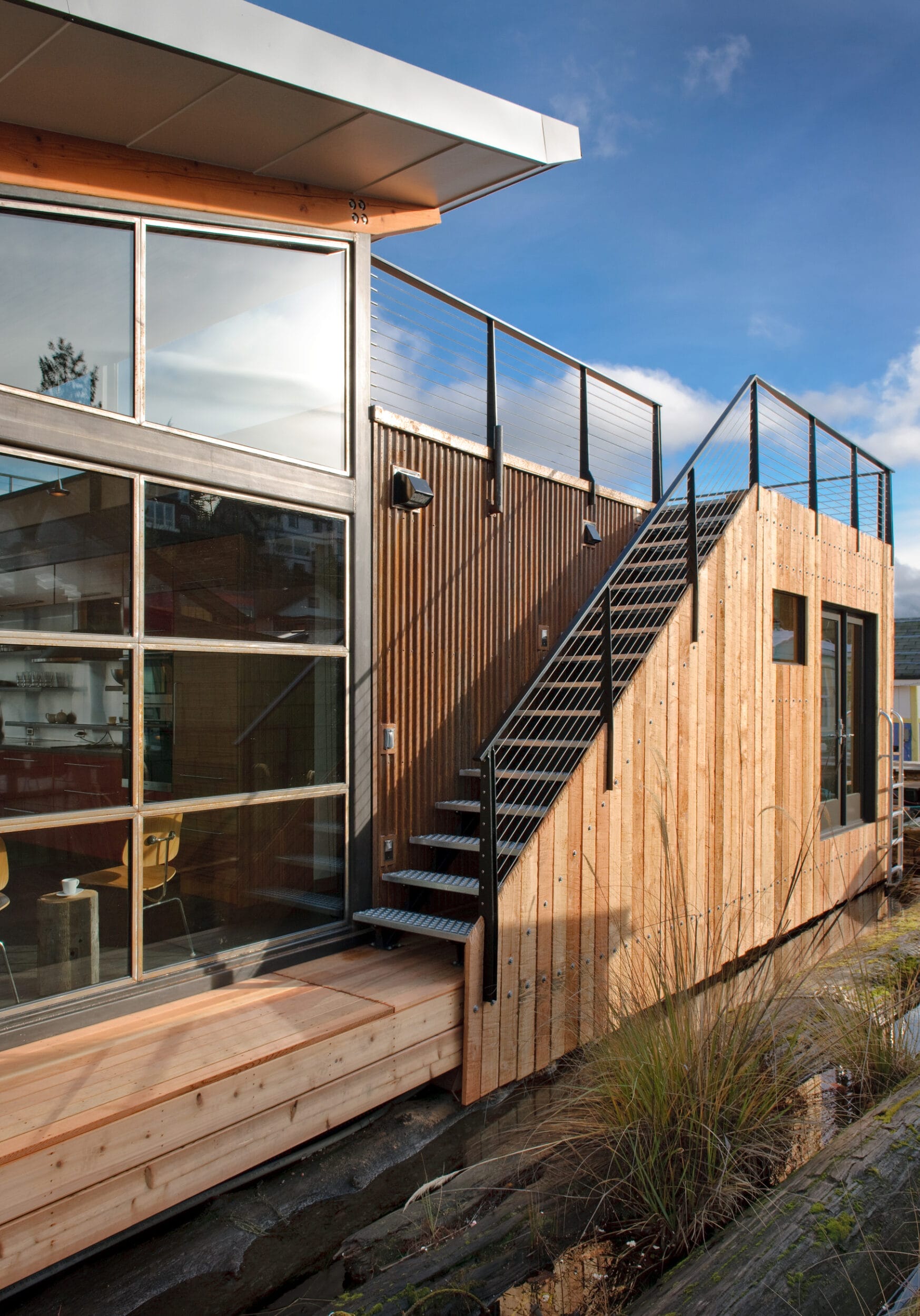 Modern wooden building exterior with large windows and an outdoor metal staircase. Upper deck has railing; grasses grow near the base. Sky is clear and blue.