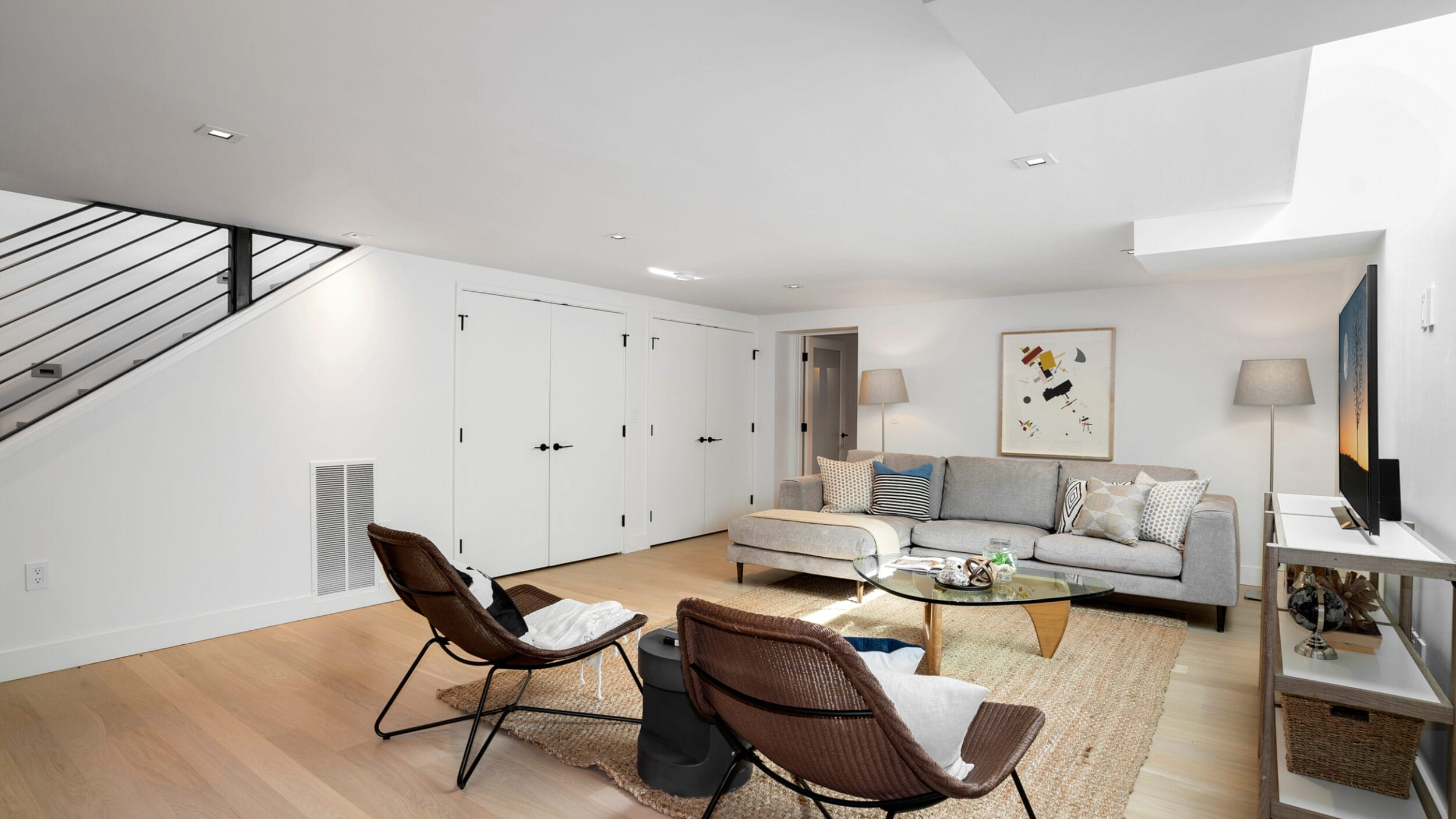 Modern living room with a gray sofa, two brown chairs, a glass coffee table, and decorative artwork. Light wood flooring complements the minimalist decor. White doors under staircase.