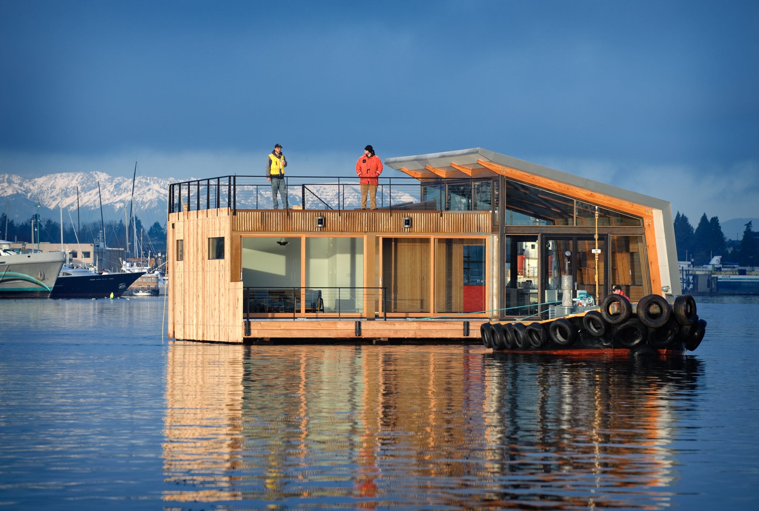 Modern houseboat on a calm body of water, with two people on the upper deck. The structure has glass walls and a sloped roof. Snow-capped mountains are visible in the background.