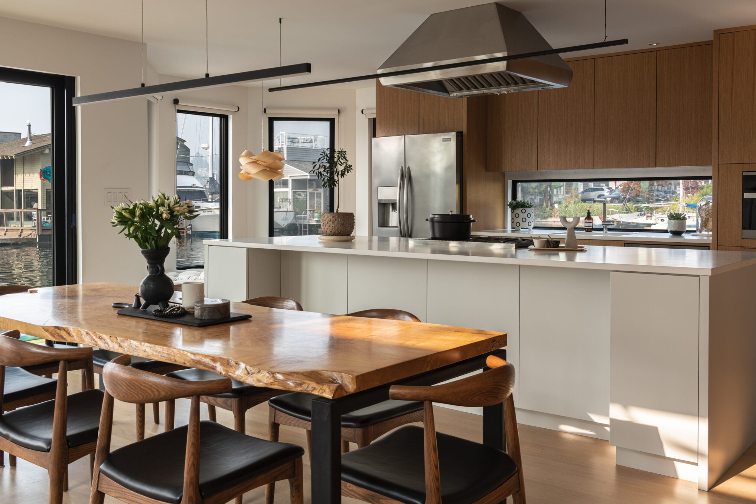 Modern kitchen with a wooden dining table, six chairs, and a kitchen island. Stainless steel appliances, overhead vent, and a light fixture above. Large windows reveal an outdoor view.