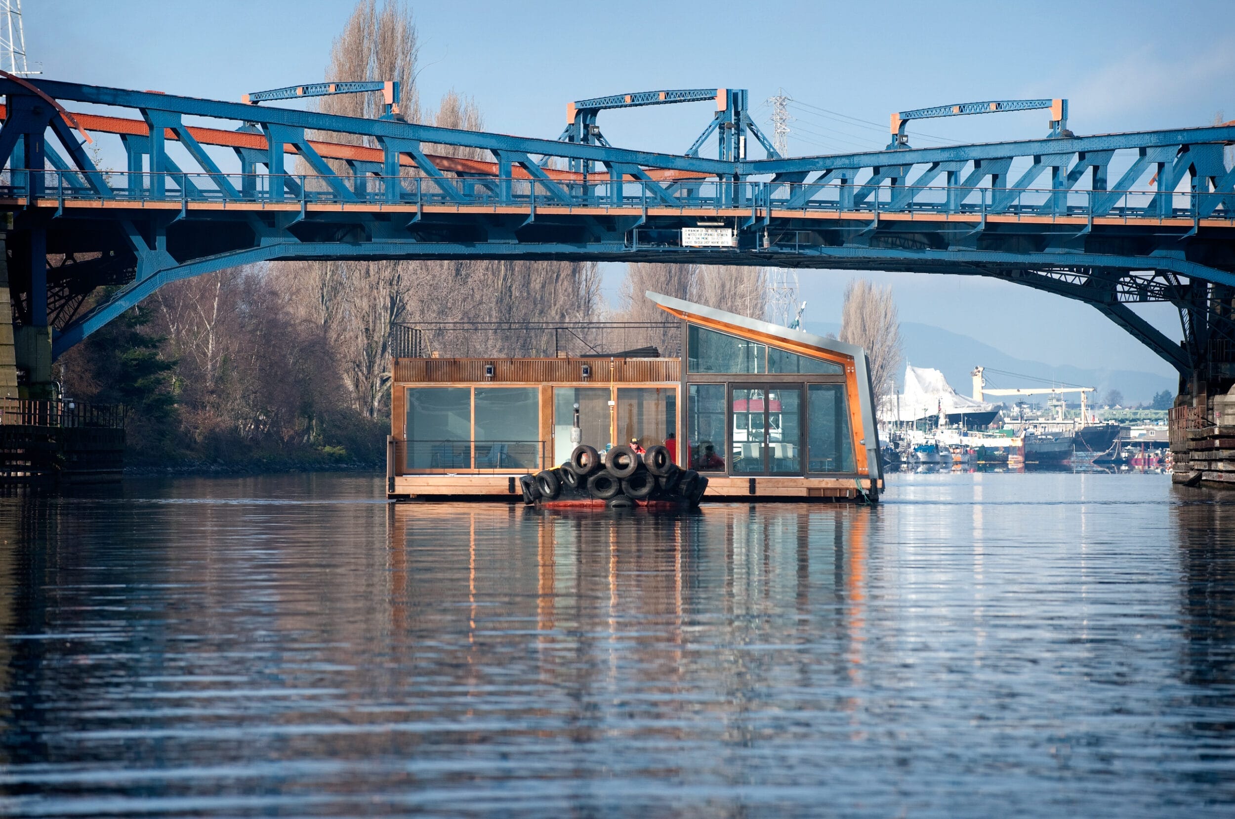 Houseboat on calm water under a blue steel bridge, with trees and industrial structures in the background on a clear day.