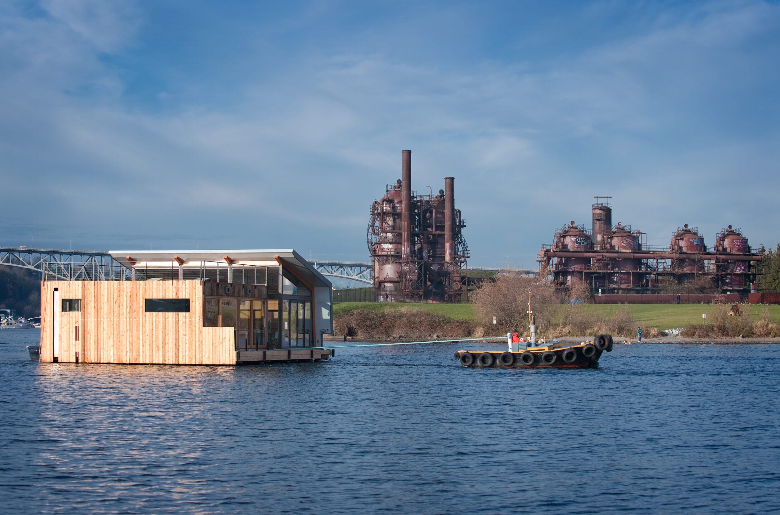 A small boat tows a wooden floating house across a body of water, with an industrial factory and a bridge visible in the background under a blue sky.