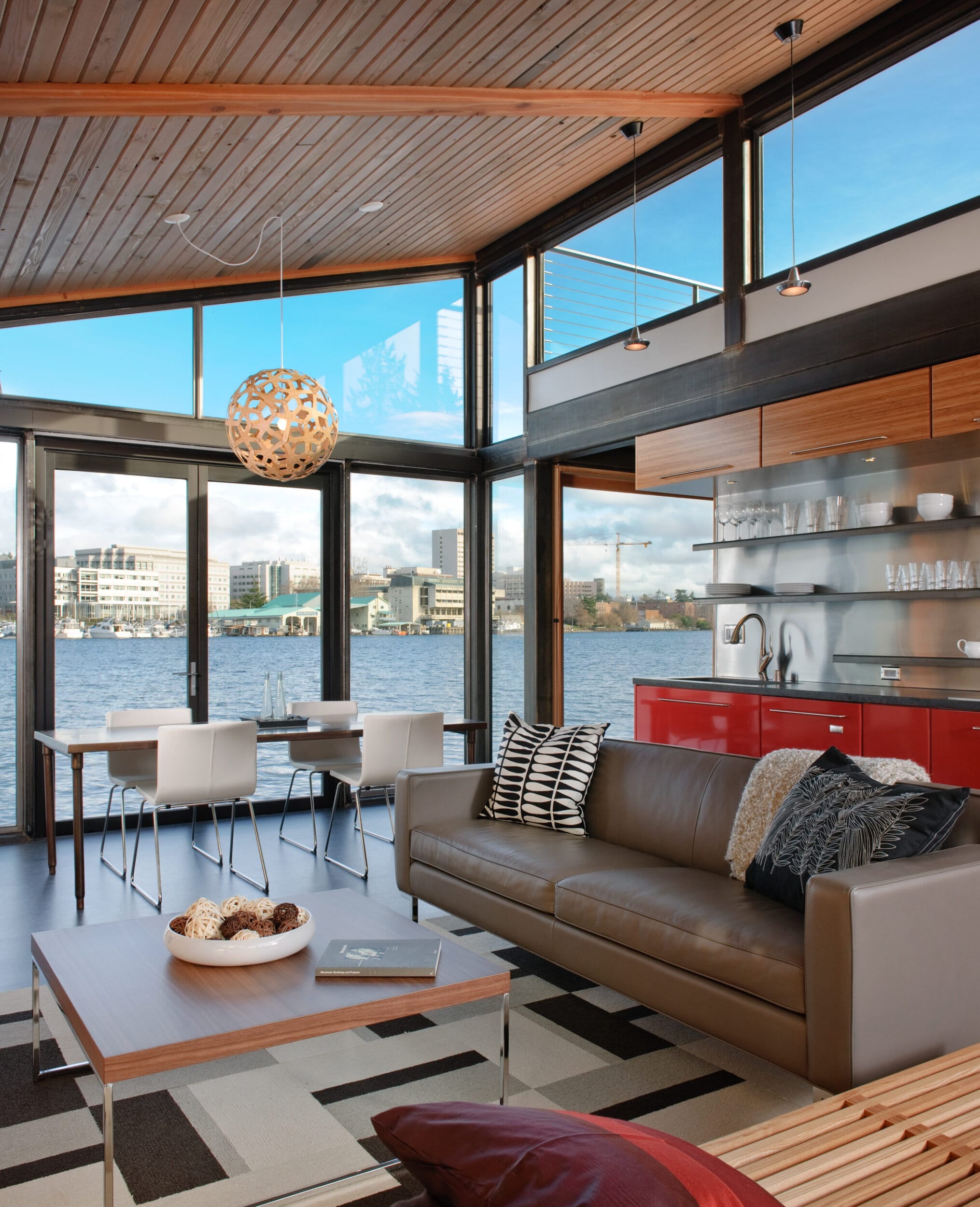 Modern living room with large windows overlooking a body of water. Features a gray sofa, wooden ceiling, red kitchen cabinets, and a dining table with white chairs. A round light fixture hangs above.