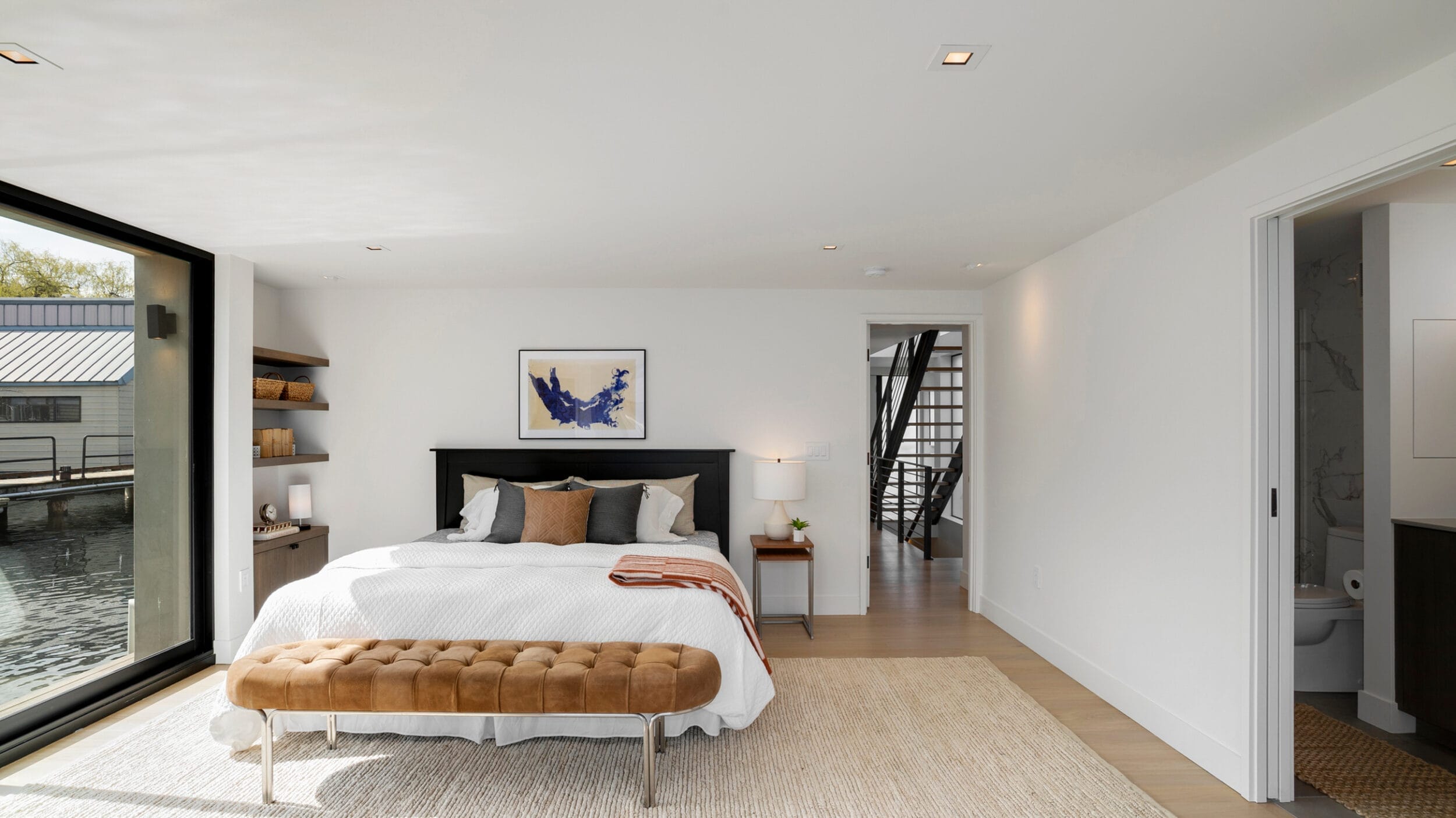 Modern bedroom with a large window overlooking water, featuring a bed with white bedding, a brown bench, shelves, a nightstand with a lamp, and an open door to a bathroom.