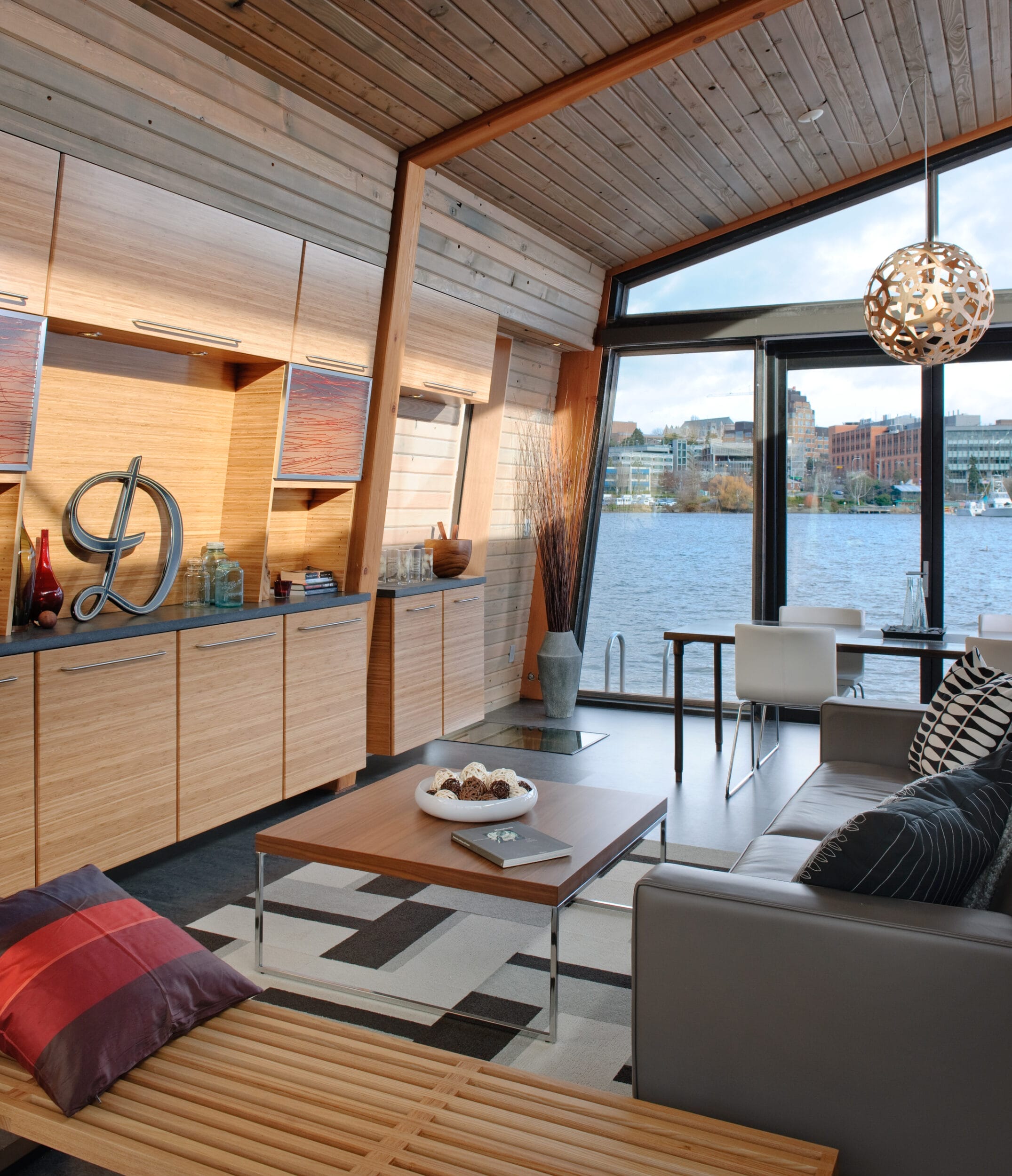 Modern living room with wooden accents, large windows overlooking a lake, a geometric rug, pillows on a bench, a coffee table, and a decorative hanging light fixture.