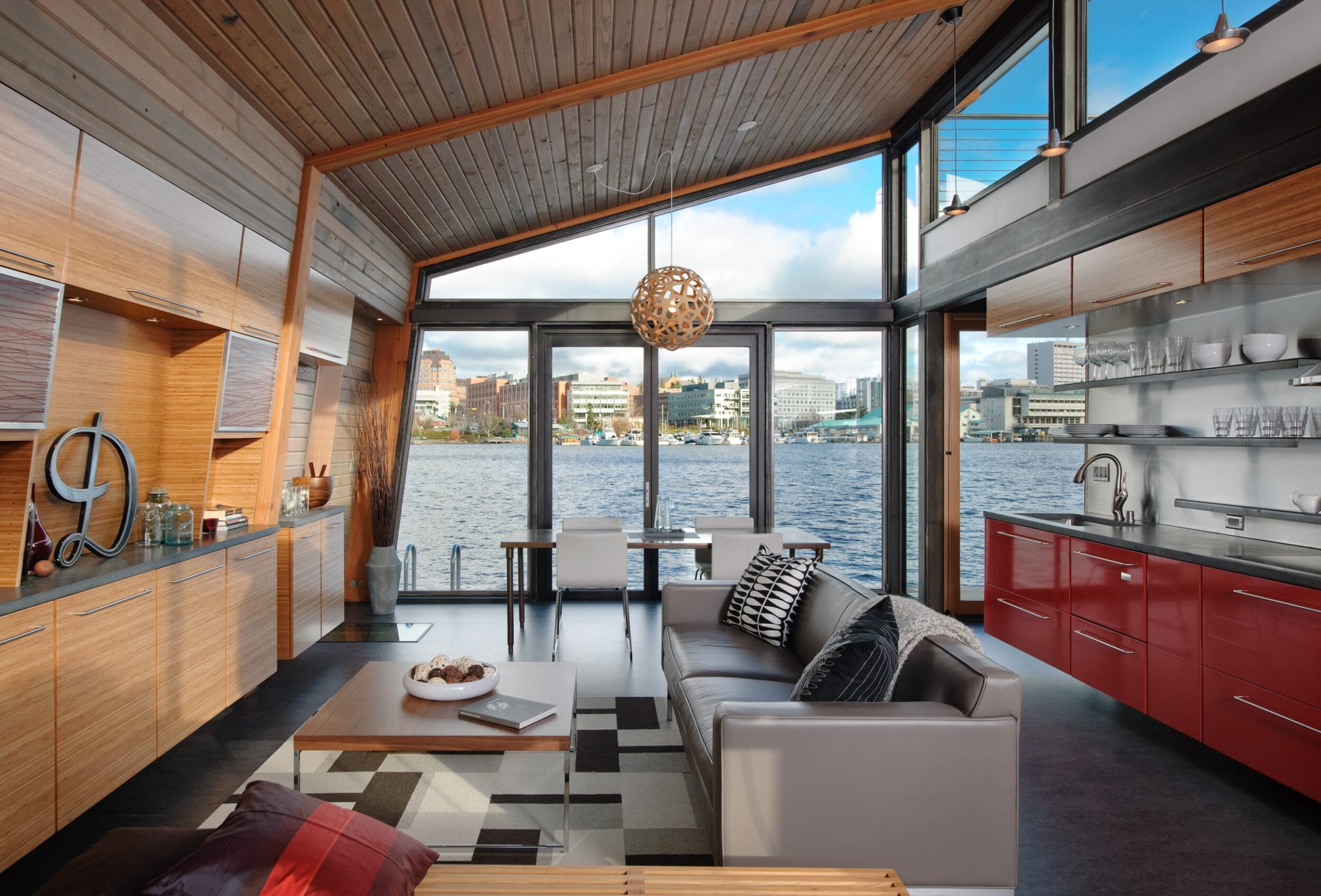 Modern living room with wooden ceiling and red cabinets, overlooking a large body of water through expansive windows. White sofa and dining table are present.