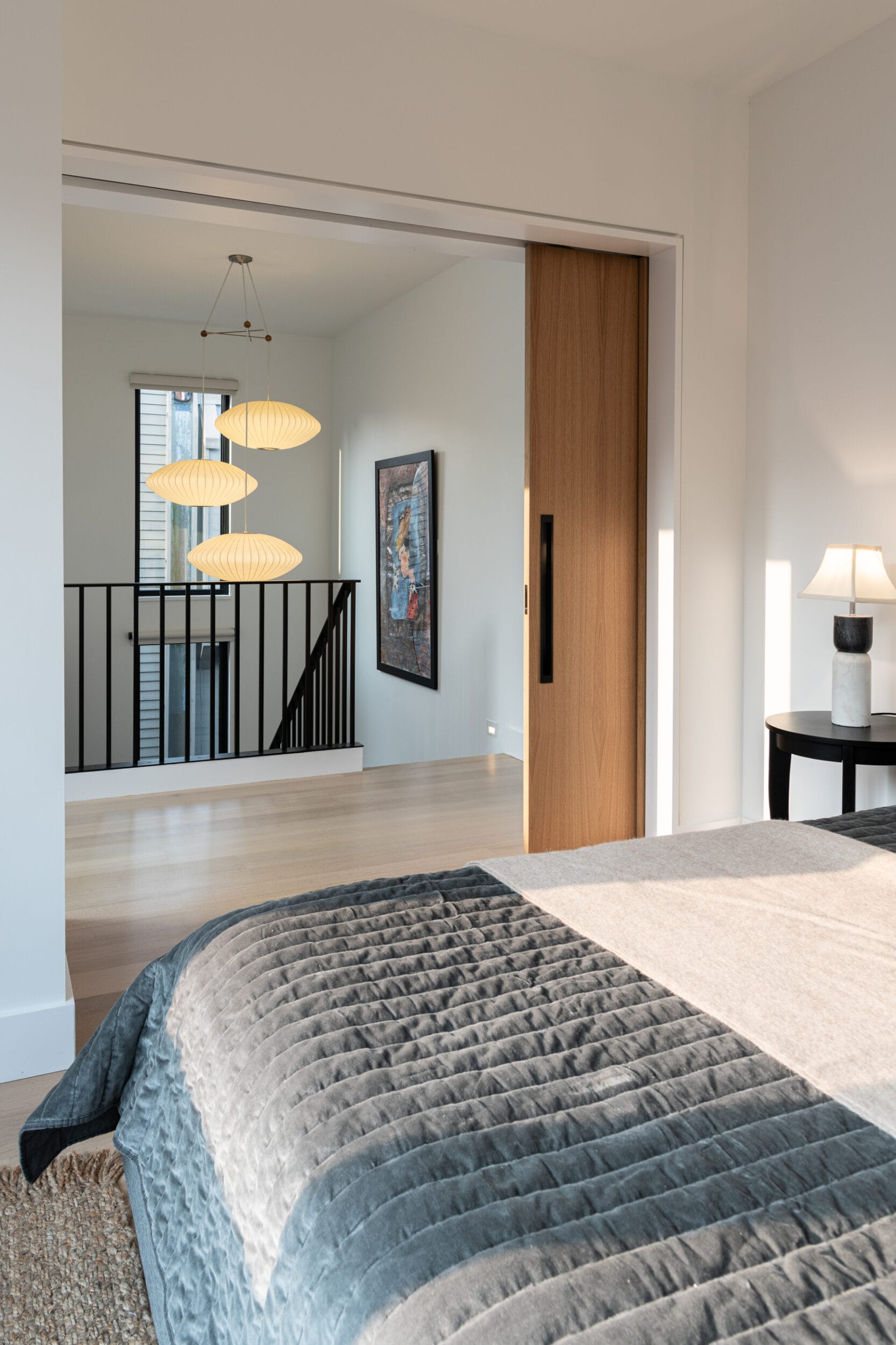 Bedroom with a neatly made bed, gray bedding, and a lamp on a side table. Sliding wooden door opens to a hallway with hanging light fixtures and a framed artwork on the wall.