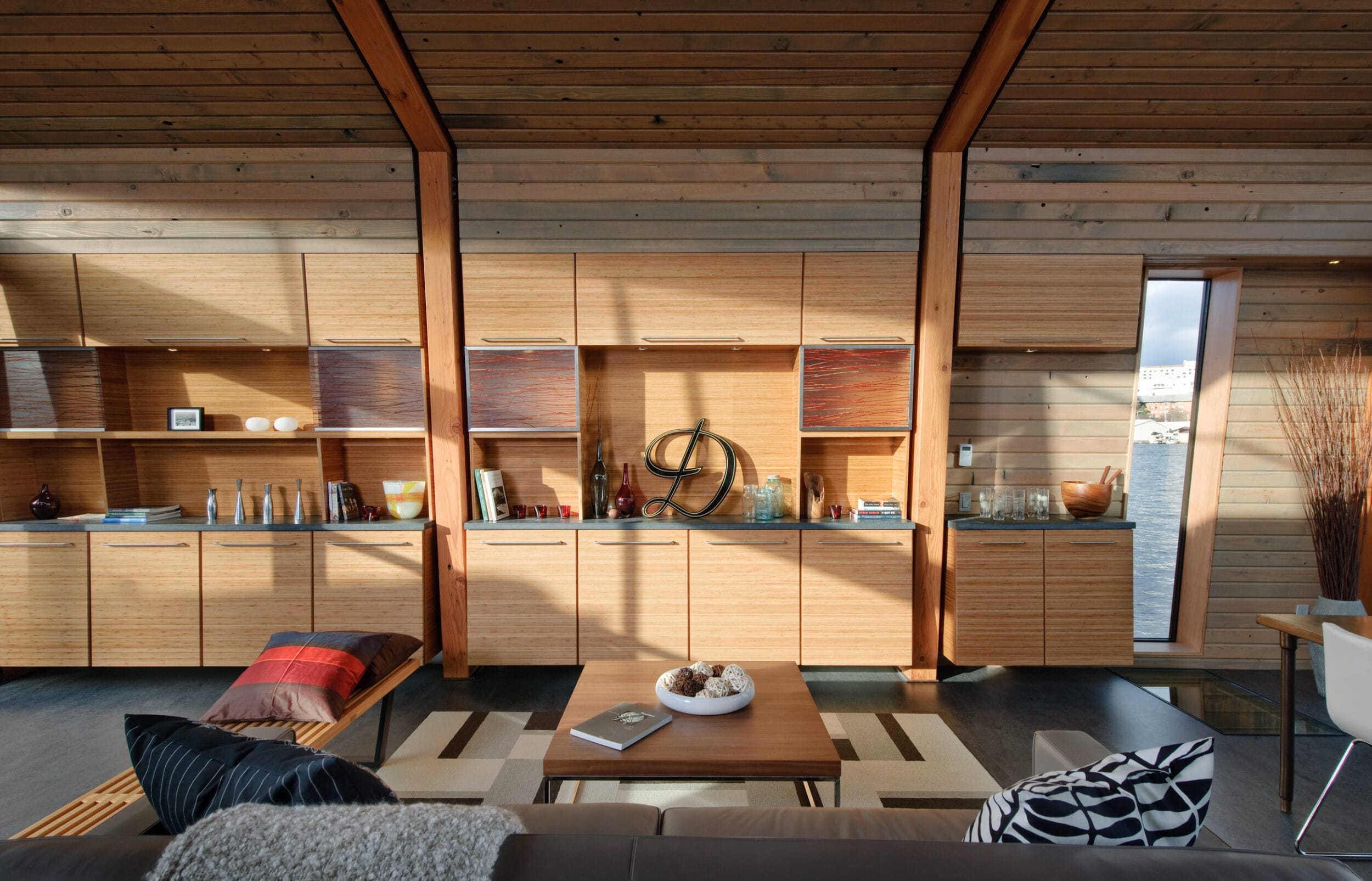 Modern wooden interior with large windows, shelves, and cabinets. A sofa with cushions faces a central wood table. Dining area visible to the right.