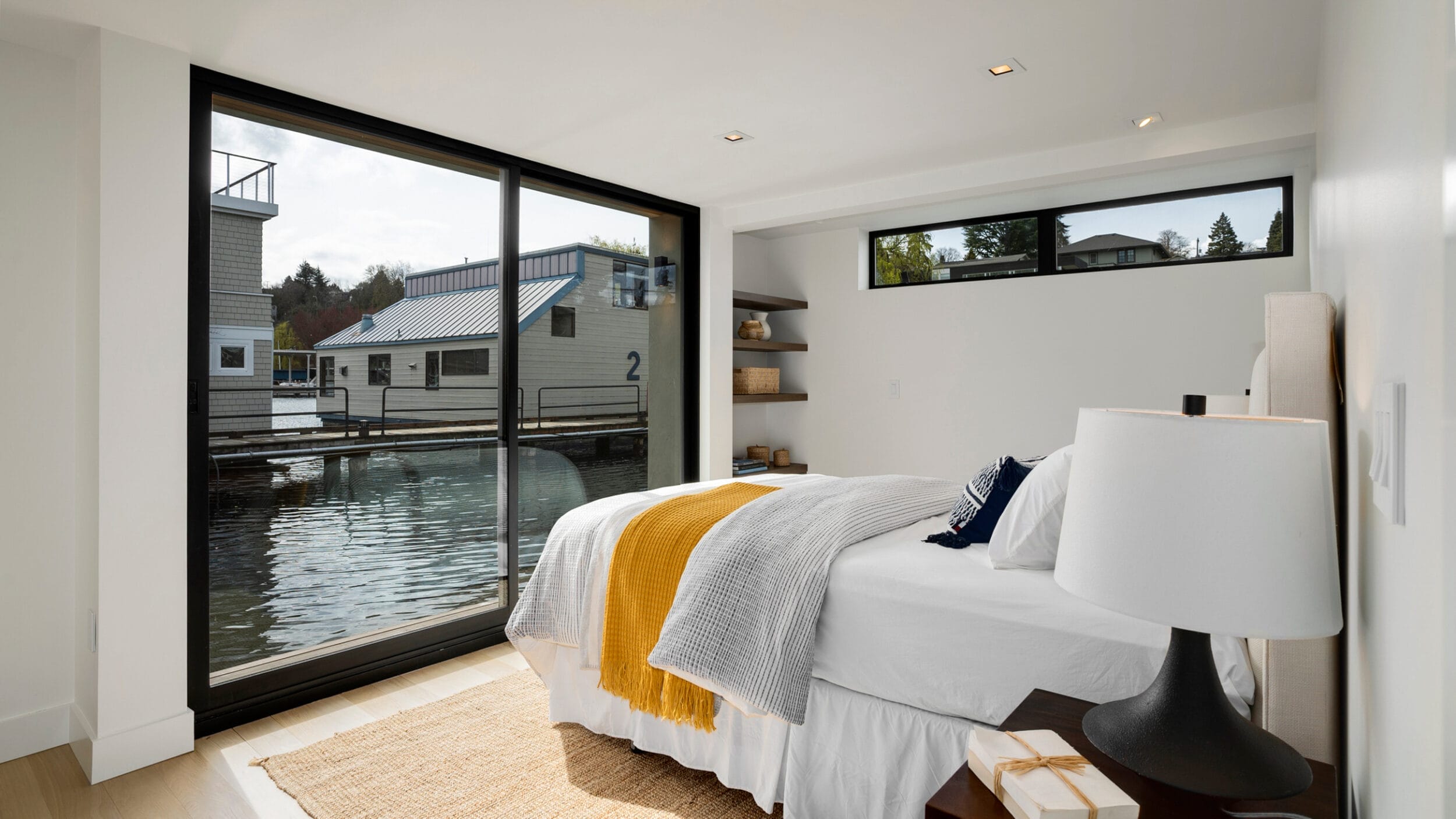 Modern bedroom with sliding glass doors overlooking a waterfront. Room features a bed with white and yellow bedding, a lamp on the nightstand, and built-in shelves.