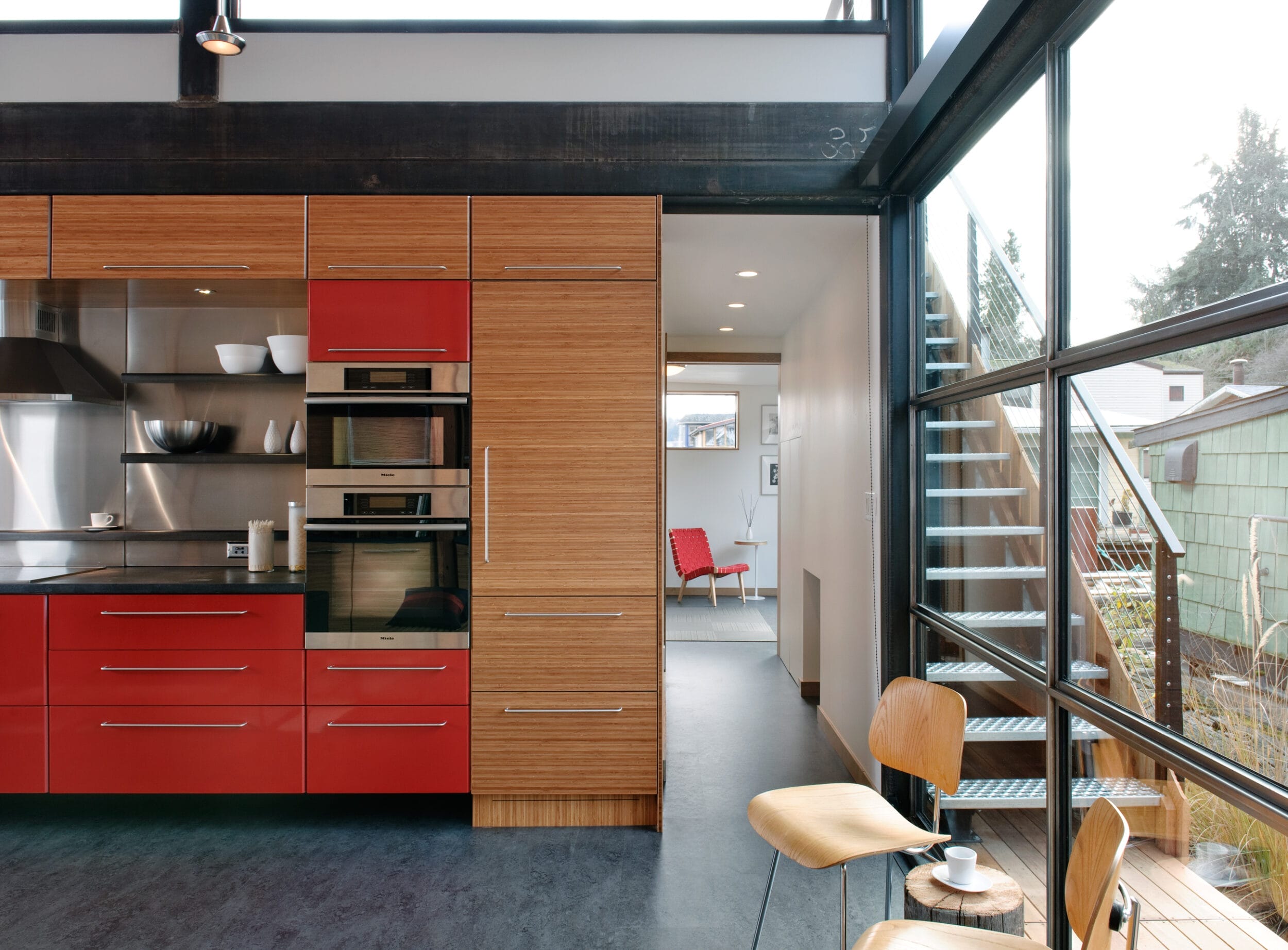 Modern kitchen with red and wood cabinetry, built-in oven, and large glass windows. Stairs lead outside, and a view of a bright room with a red chair is visible.