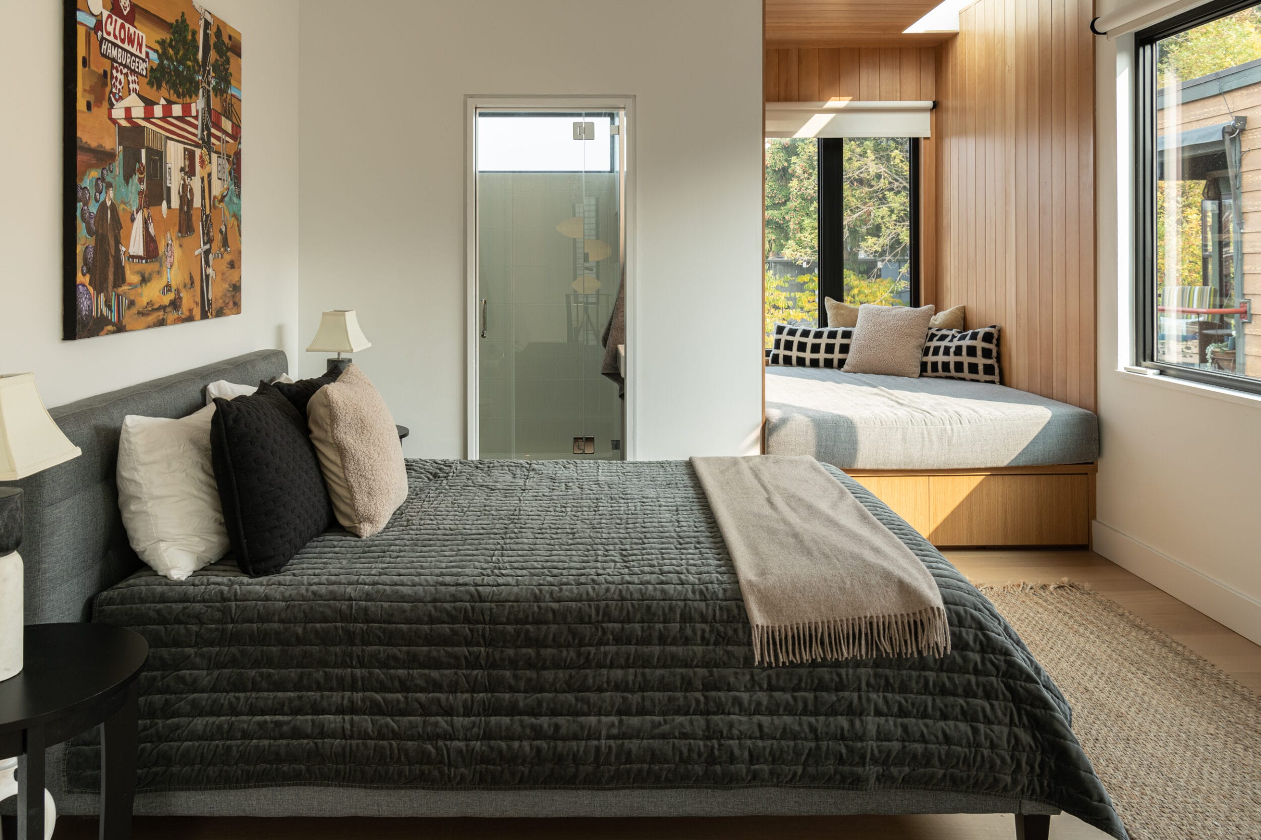 A bedroom with a large bed and black pillows in the foreground, and a smaller bed with a checkered pillow in an alcove by a window in the background. A painting is on the wall.