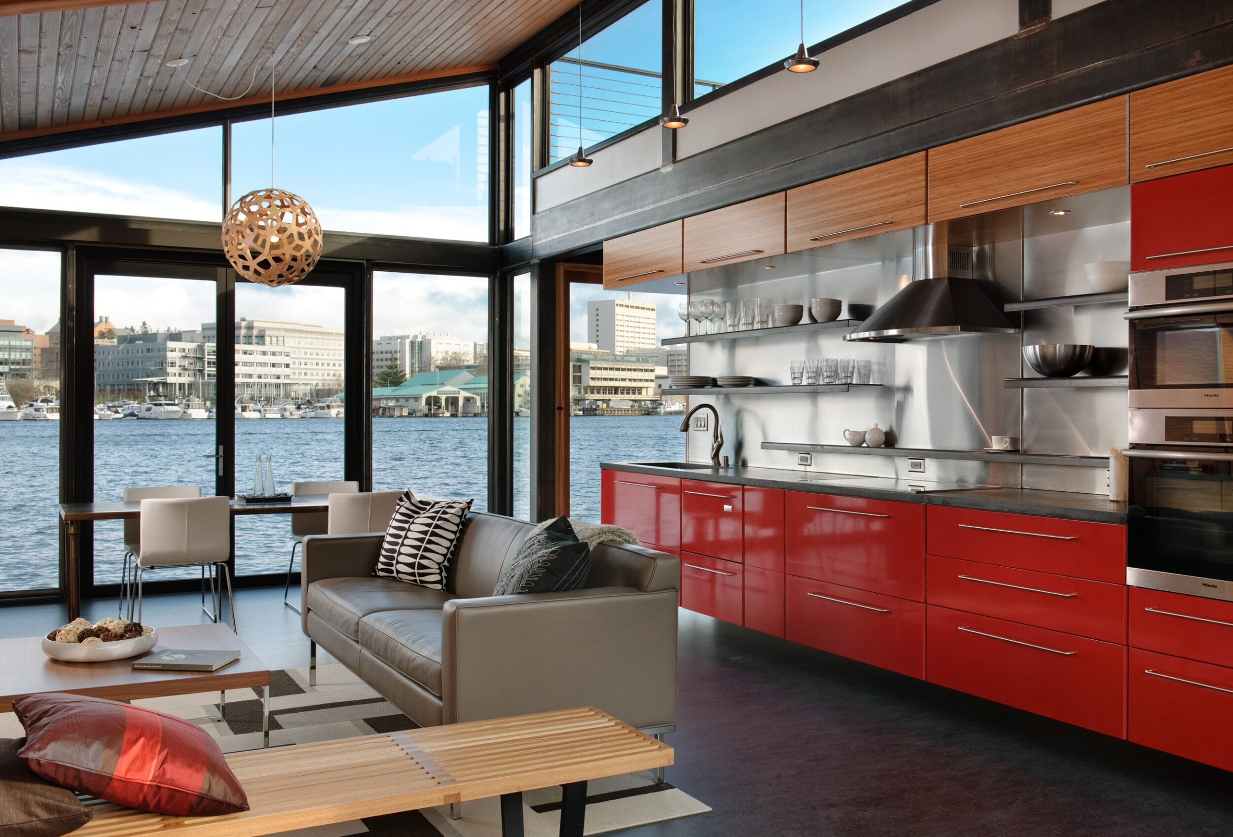 Modern kitchen and living area with red cabinets, stainless steel appliances, and large windows offering a waterside city view. Includes a sofa, coffee table, and dining set.