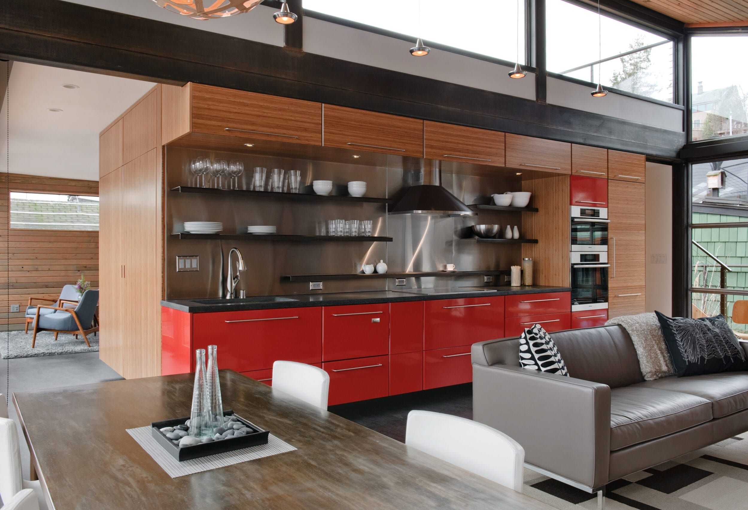 Modern kitchen with red cabinets, stainless steel appliances, and open shelving. Adjacent dining table and gray couch in an open-concept living area with large windows and wooden accents.