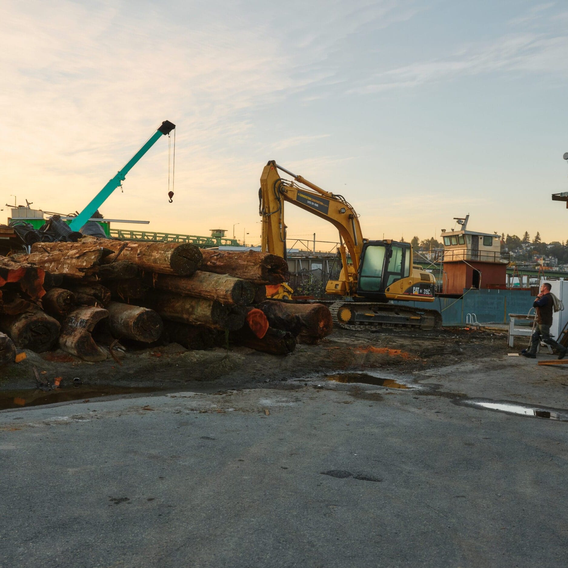 Large logs are stacked on the ground near construction equipment, including an excavator and a crane, at an industrial worksite with buildings in the background—ideal for a Seattle Floating Homes project by an expert floating home builder.