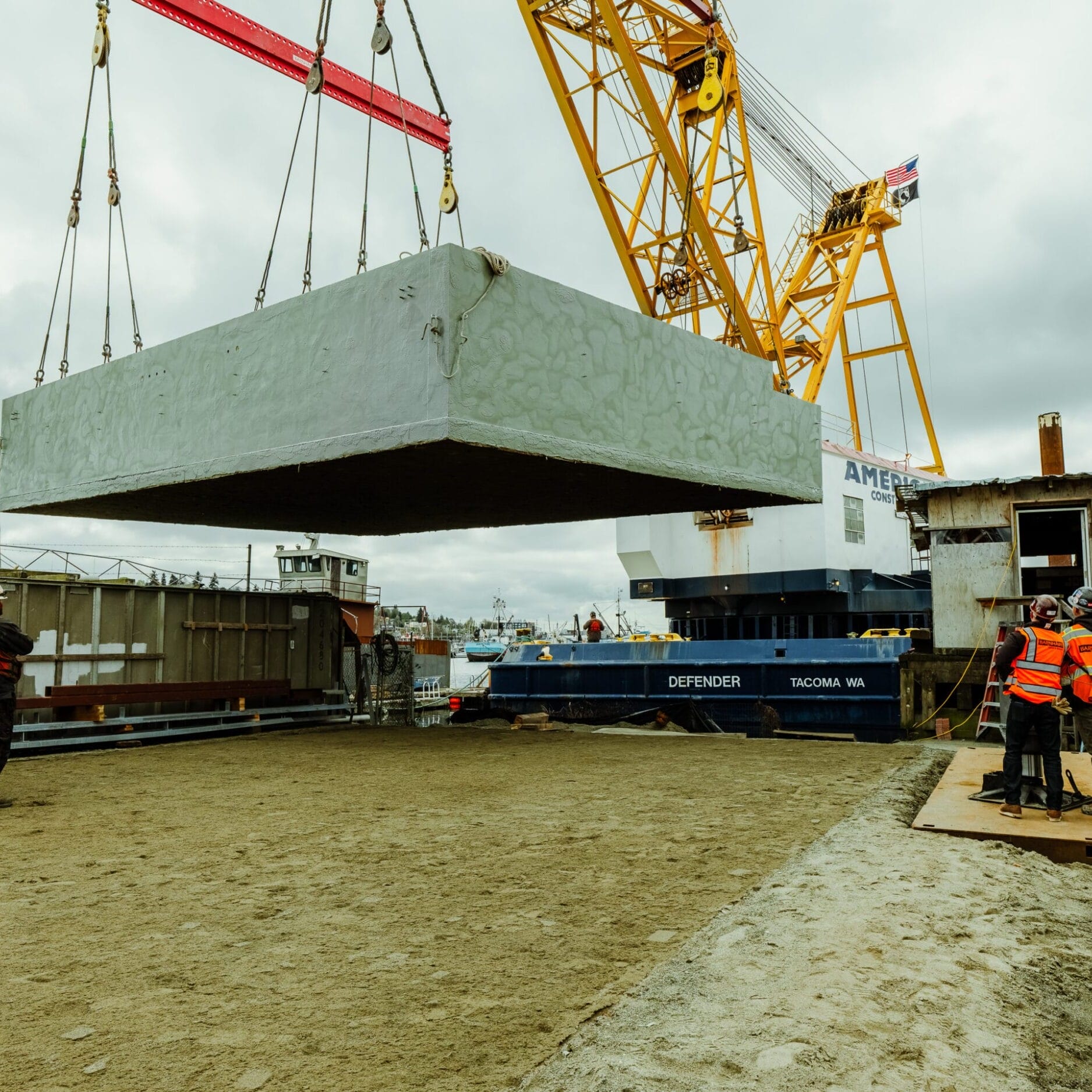 A large concrete slab is being lifted by a crane as workers in safety gear oversee the operation on a construction site near a docked vessel, guided by the expertise of a Seattle Floating Homes contractor.
