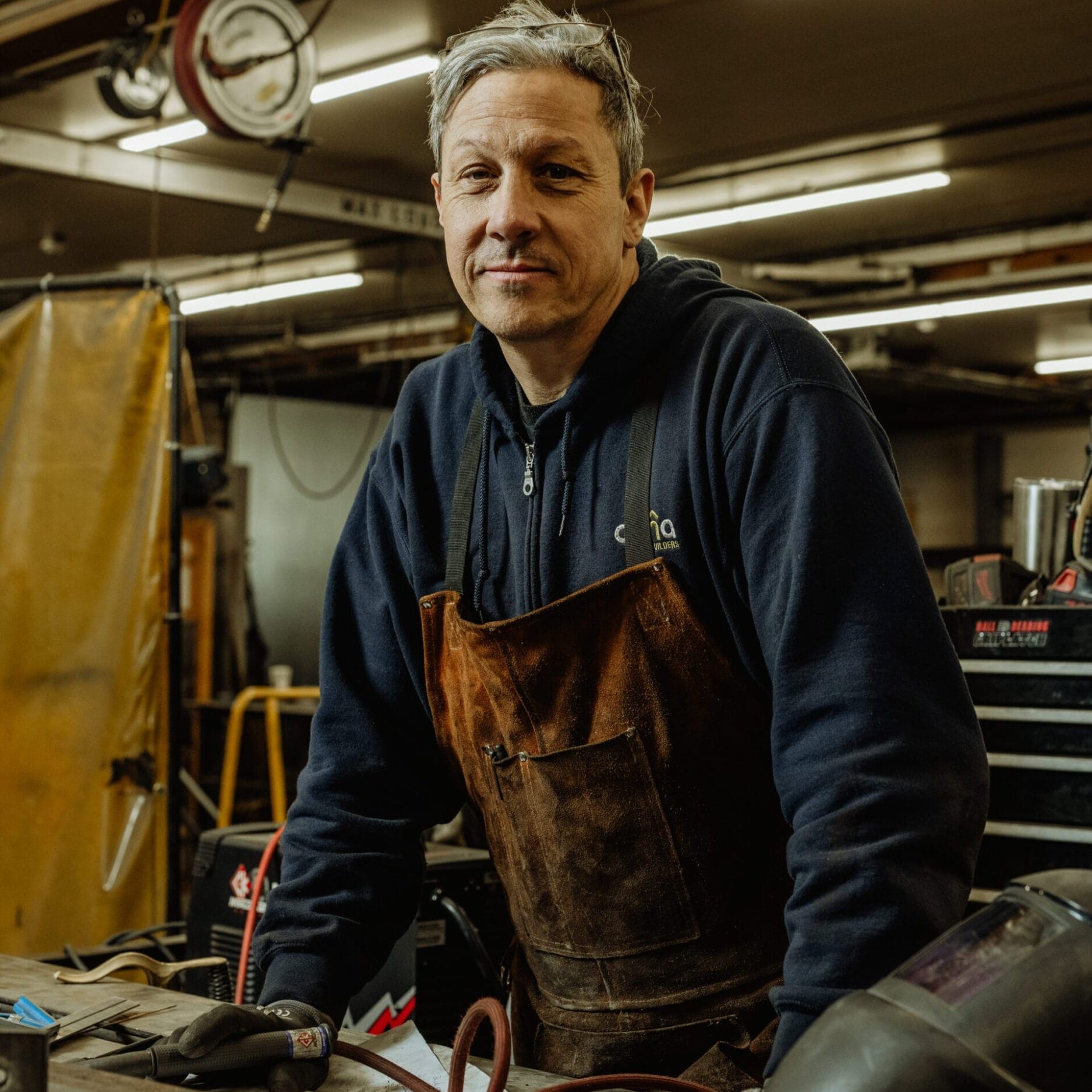 A man wearing a brown apron and gloves stands at a workbench in the Dyna Metal Shop Seattle workshop, with tools and equipment visible in the background.