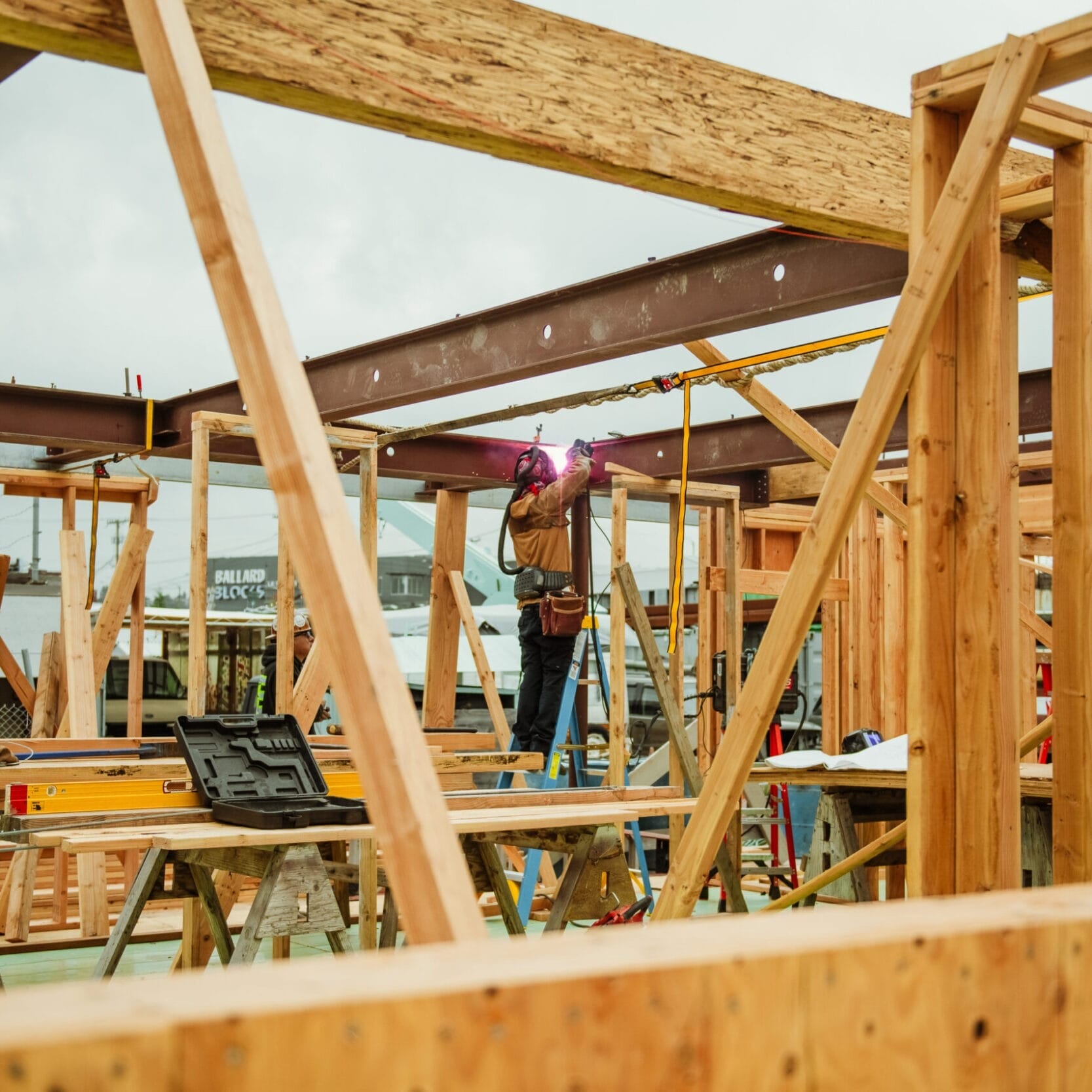 A construction worker stands on a ladder welding a metal beam in a wooden building frame at an outdoor site, guided by the expertise of a Seattle Floating Homes contractor.
