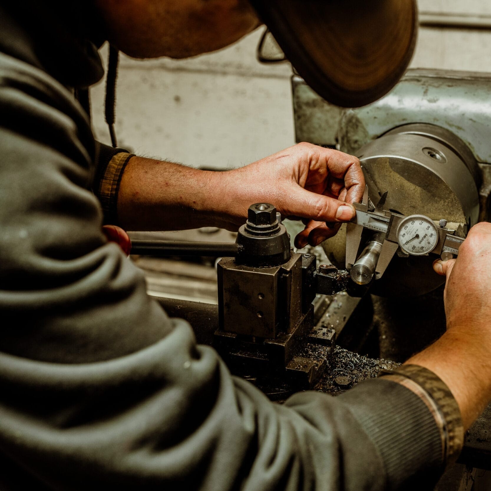 A person uses a caliper to measure a metal object on a lathe machine at Dyna Metal Shop Seattle in a busy workshop setting.