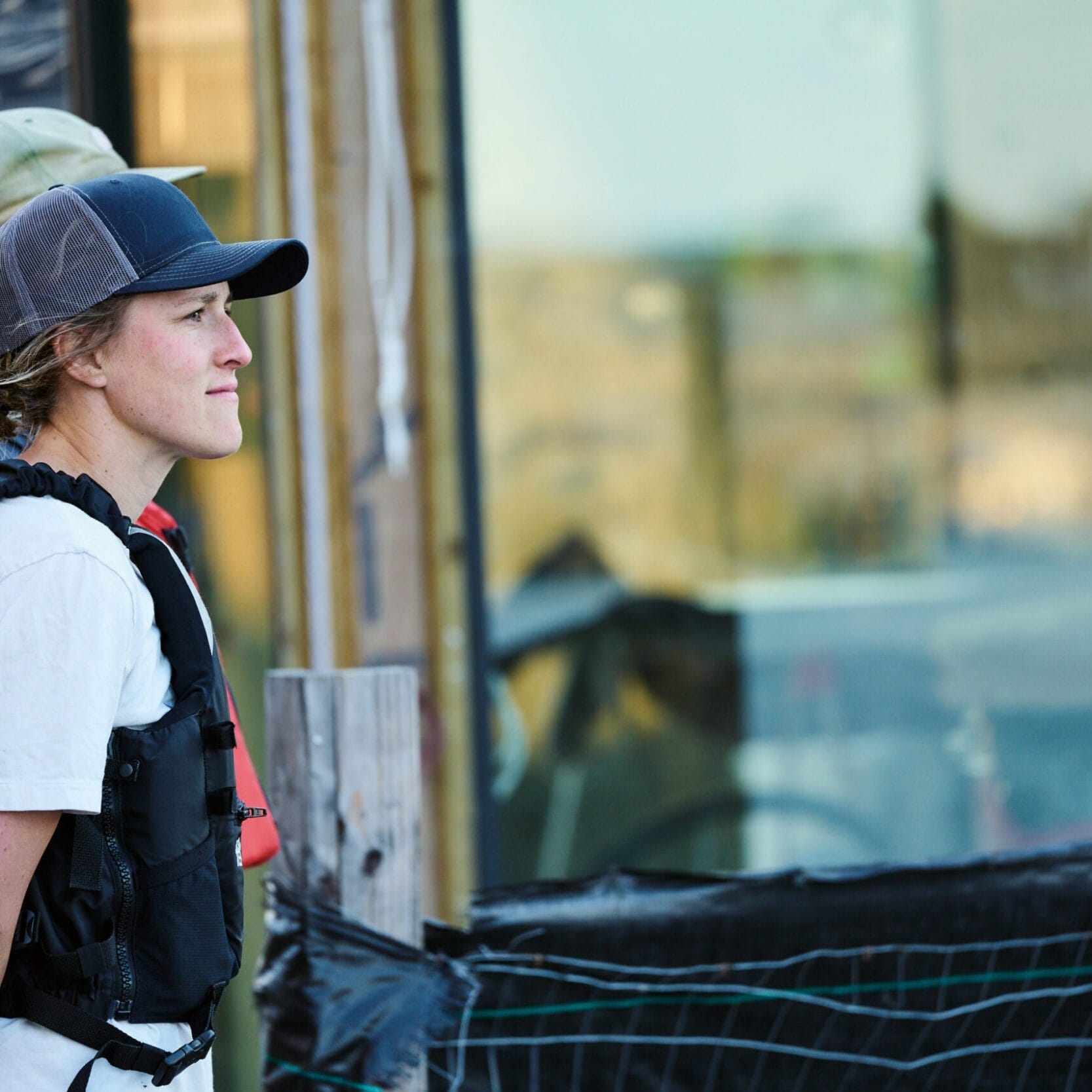 Two people wearing caps and life vests stand outdoors near a building with large windows, possibly discussing plans with a Seattle Floating Homes contractor.