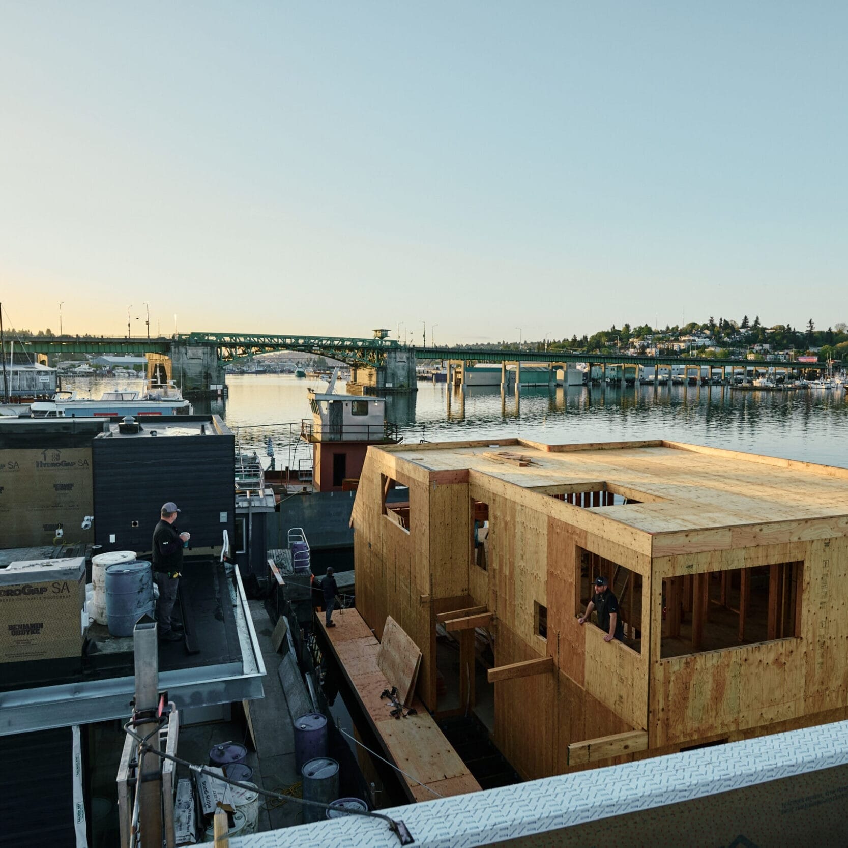 A wooden house is under construction on a dock near a body of water, with workers on site and boats and buildings visible in the background—an example of a Seattle Floating Homes contractor at work.