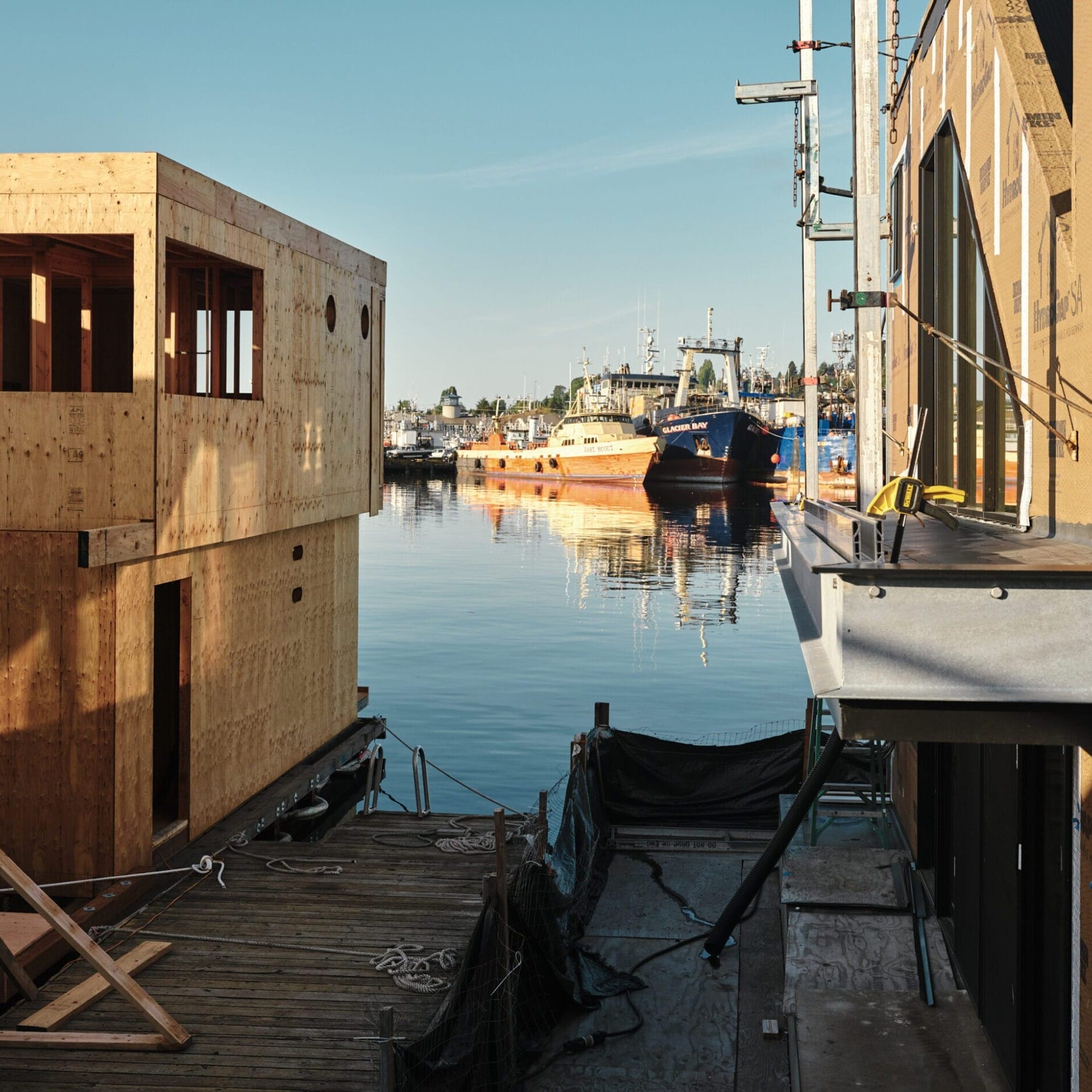 Partially constructed wooden buildings by the dock suggest the work of a skilled Floating Homes Contractor, with calm water and several boats and ships moored in the background under a clear sky.