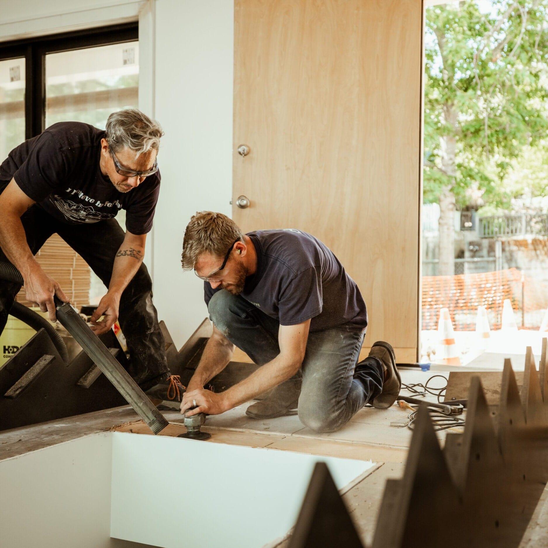 Two men from Dyna Metal Fabricators install or adjust a staircase railing indoors, using tools and kneeling on the floor, with boxes and construction materials in the background.