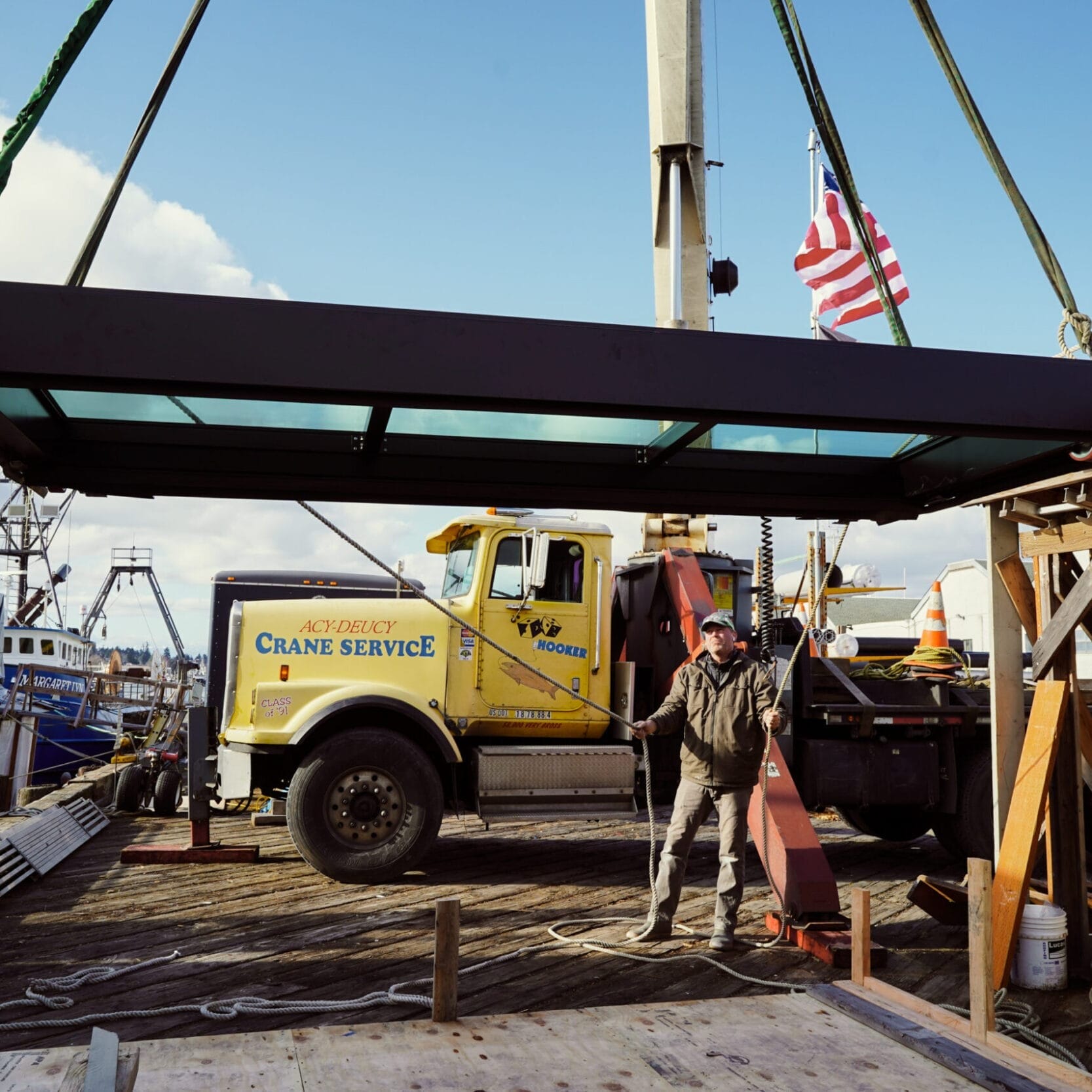 A crane lifts a large rectangular glass panel above a dock, with a worker guiding it and a yellow Crane Service truck in the background—perfect for any Seattle Floating Homes project.