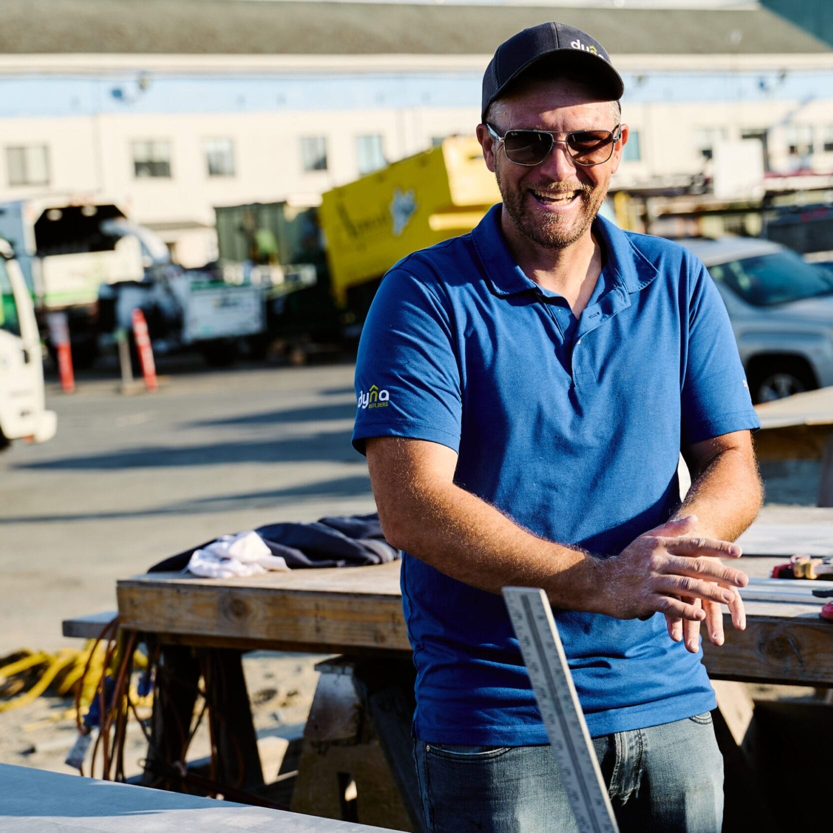 A man in a blue polo shirt and cap stands smiling next to a worktable at a construction site with trucks and tools in the background, representing a skilled Seattle Floating Homes contractor.