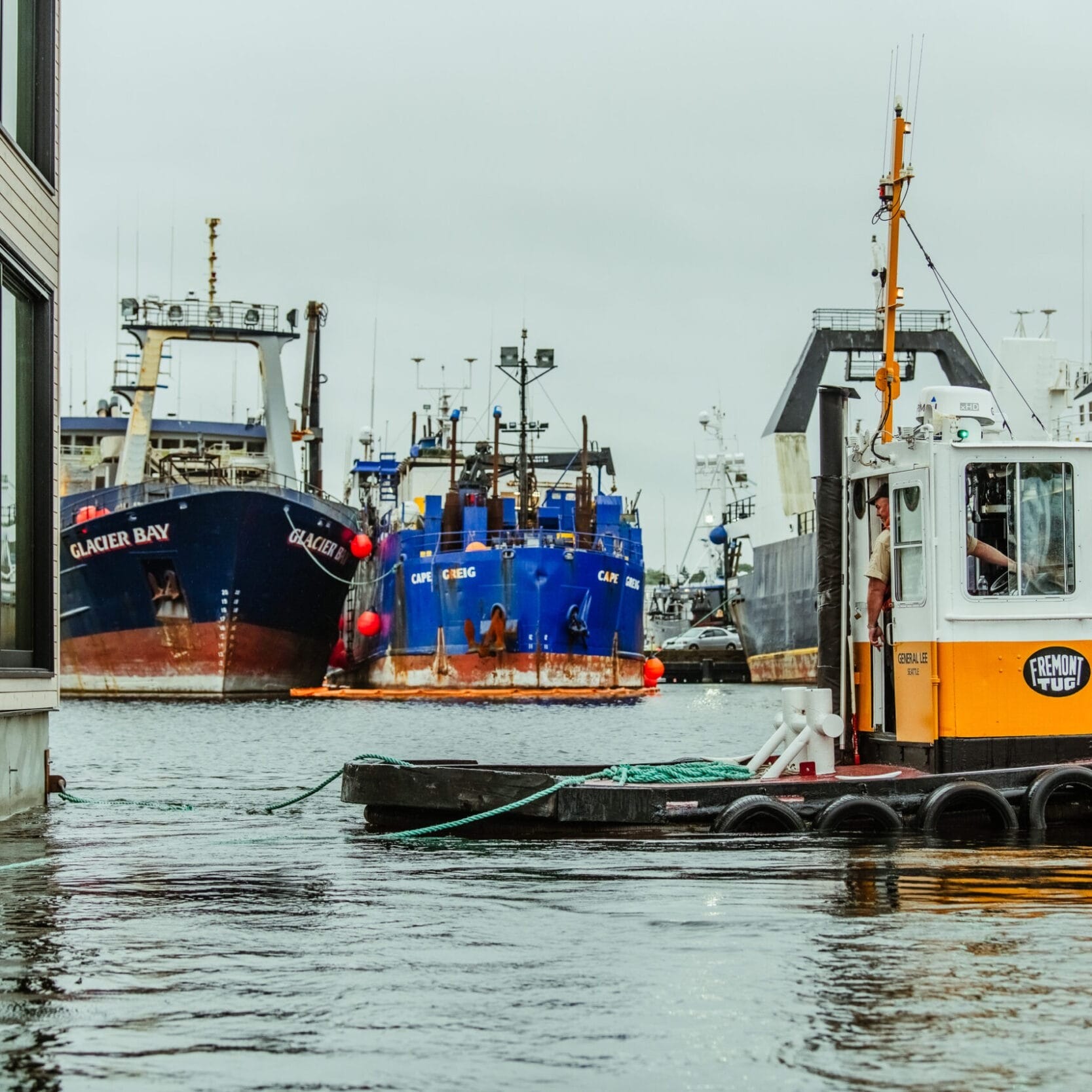 A small tugboat moves through a harbor with several large fishing vessels docked in the background on a cloudy day, near where Seattle Floating Homes and skilled floating home builders create unique waterfront residences.