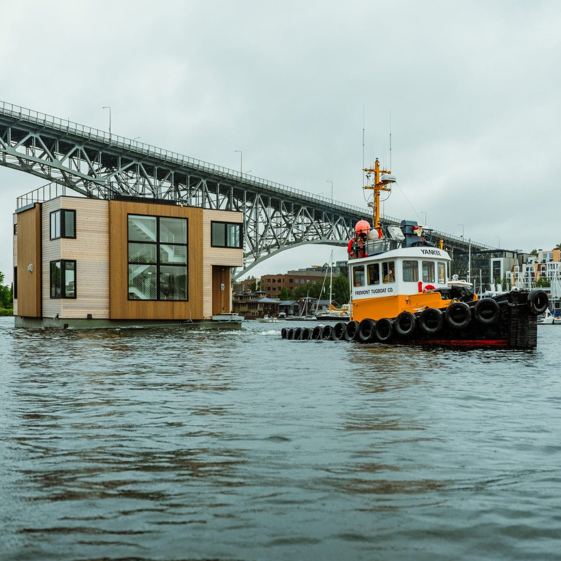 A tugboat tows a modern floating house built by a Seattle Floating Homes contractor on a river beneath a large bridge, with city buildings in the background under a cloudy sky.